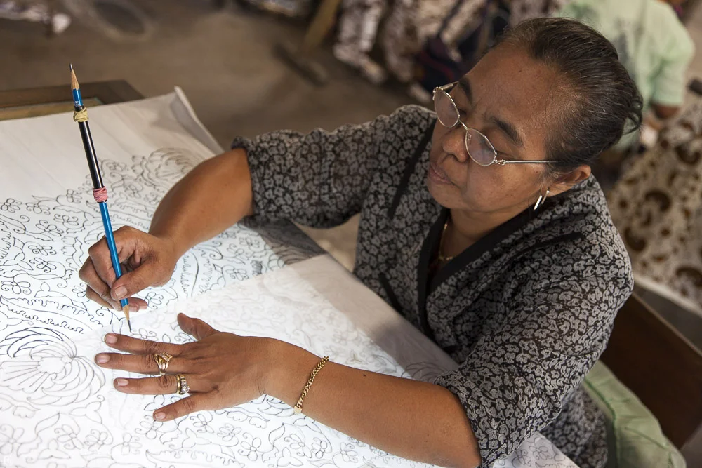 An older woman with glasses and earrings, wearing a patterned blouse, is sitting at a table drawing intricate patterns and designs on a large sheet of paper with a blue and black pen.