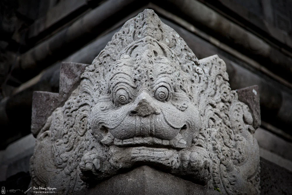 A detailed stone sculpture of a mythical creature, resembling a lion with a fierce expression, large round eyes, a prominent nose, and a wide mouth with visible teeth, located on a building or structure.