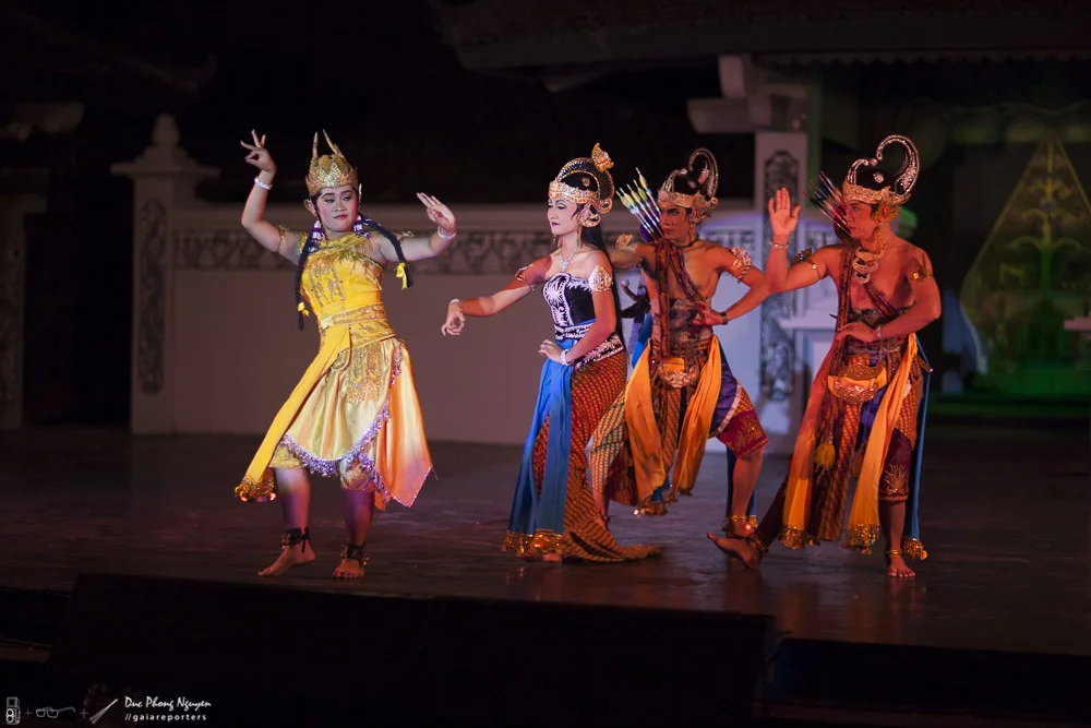 Performers in traditional Southeast Asian costumes dancing on stage under dim lighting.