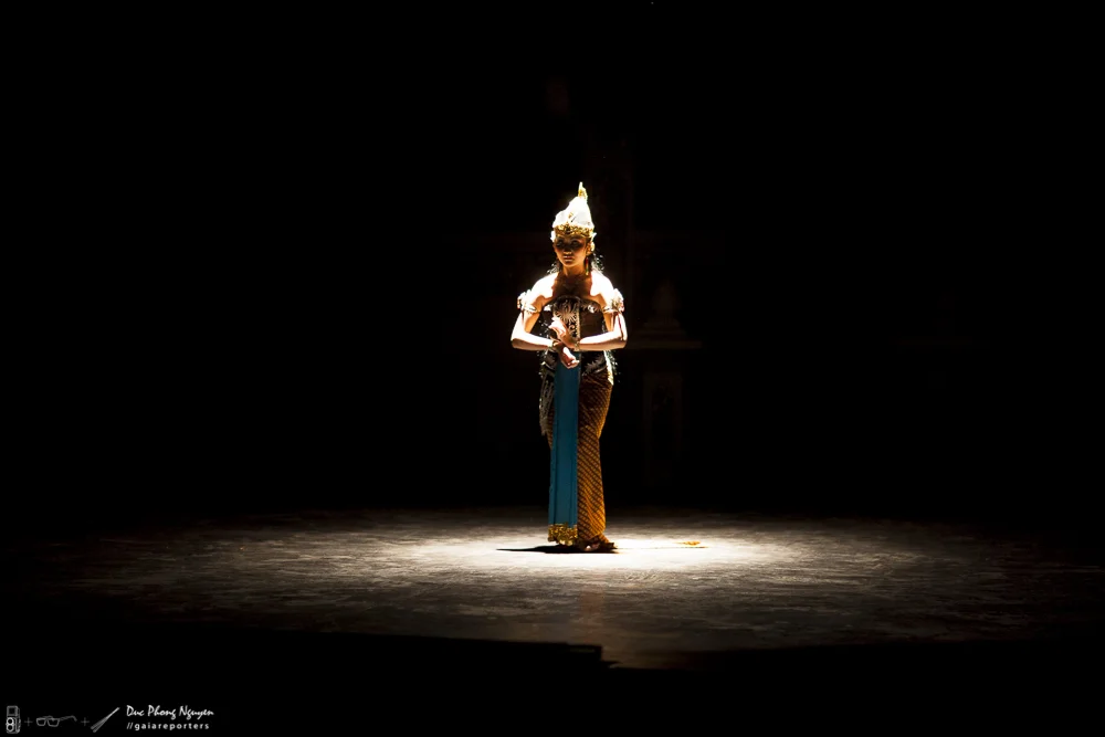 A performer dressed in traditional Southeast Asian attire standing alone on a brightly lit stage against a dark background.
