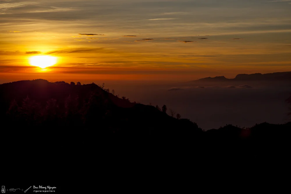 Sunset over mountains with a vibrant orange and yellow sky, some clouds, and silhouettes of hills and trees.
