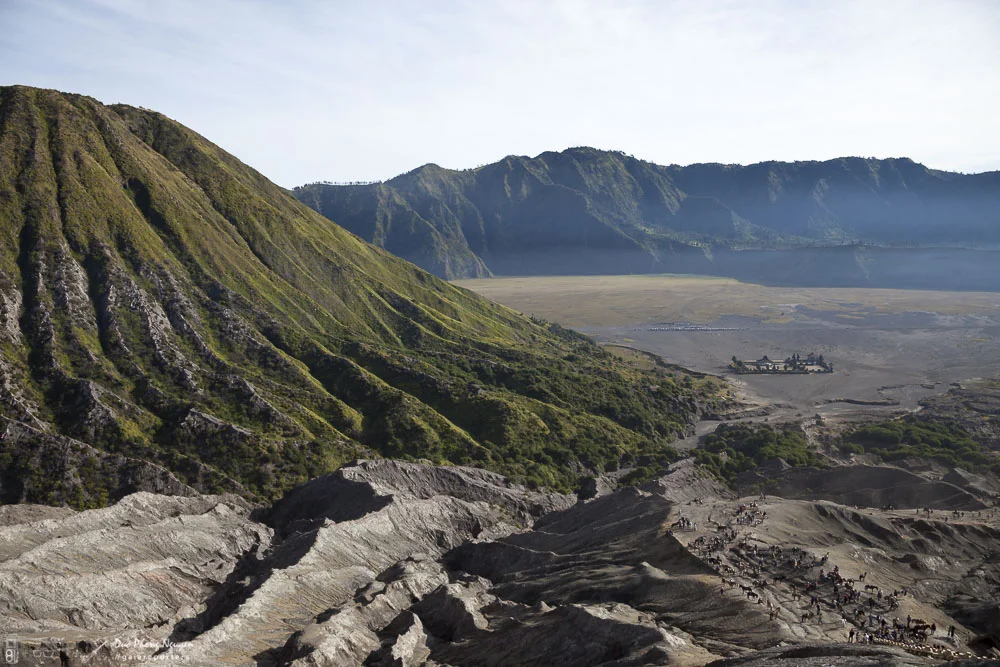 Volcanic crater with steep green slopes, a vast flat area, and a small building complex in the distance, with scattered visitors or tourists in the foreground.