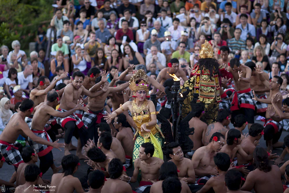 A traditional cultural dance with many performers and an audience in the background. The dancers are in colorful costumes, some with headpieces, and appear to be performing in an outdoor setting.