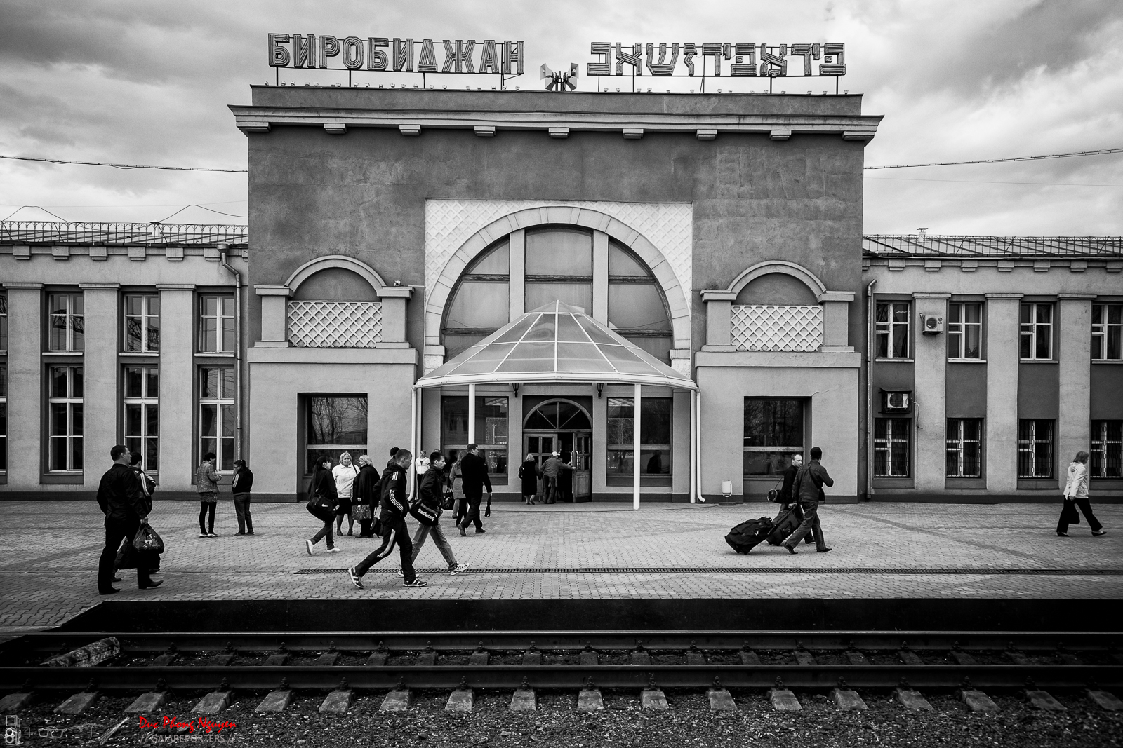 People walking and waiting outside a train station in front of a building with large windows, some with luggage, under cloudy sky.