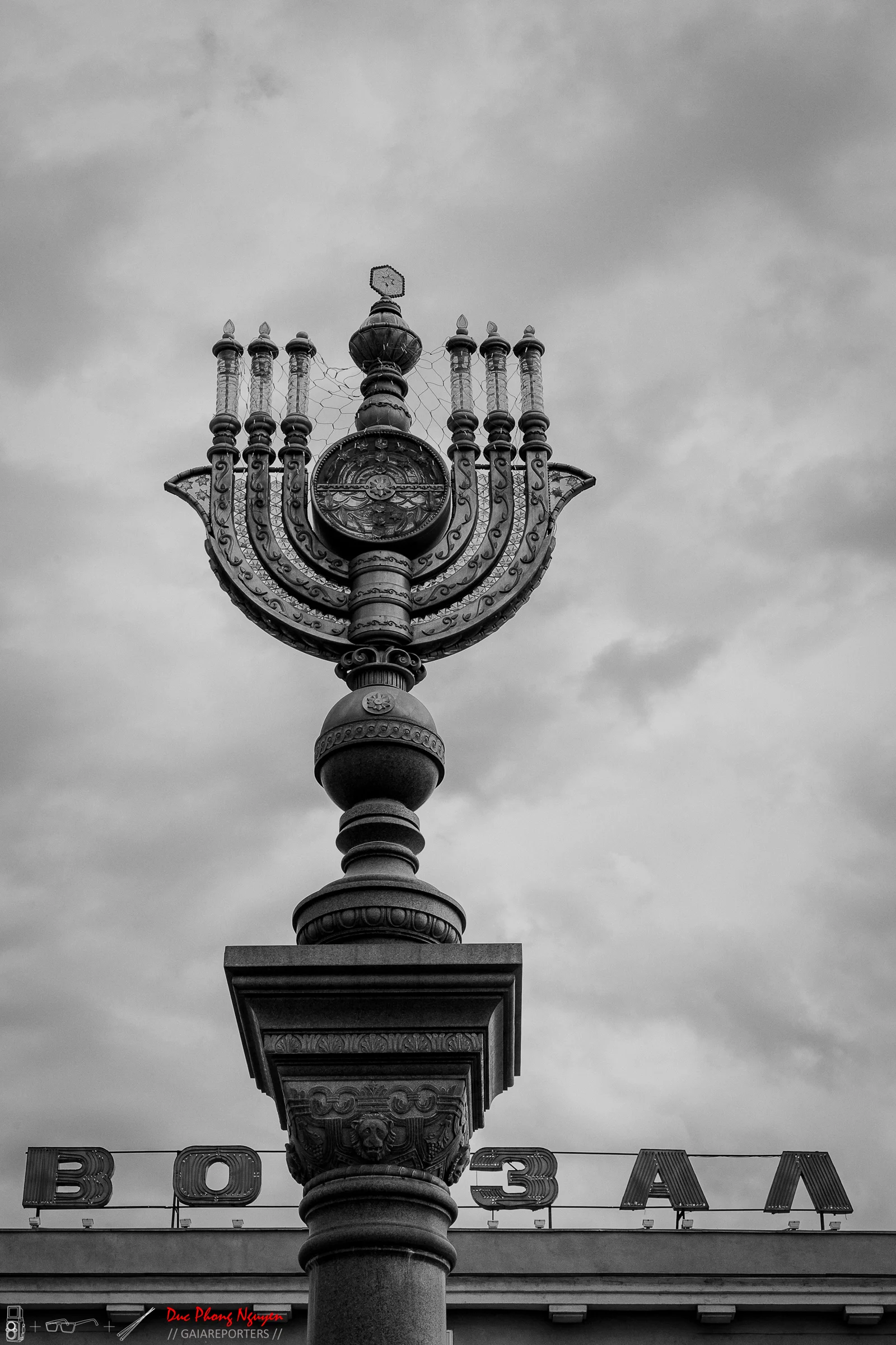 Black and white photo of a vintage menorah atop a decorative pillar in front of a building with the word "БОЛЗ" on its sign, under an overcast sky.
