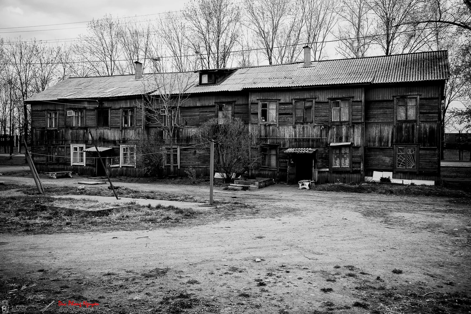 Black and white photo of an old, weathered wooden building with multiple windows and a corrugated metal roof. Bare trees are behind the building, and there are a few small structures and a dirt road in the foreground.