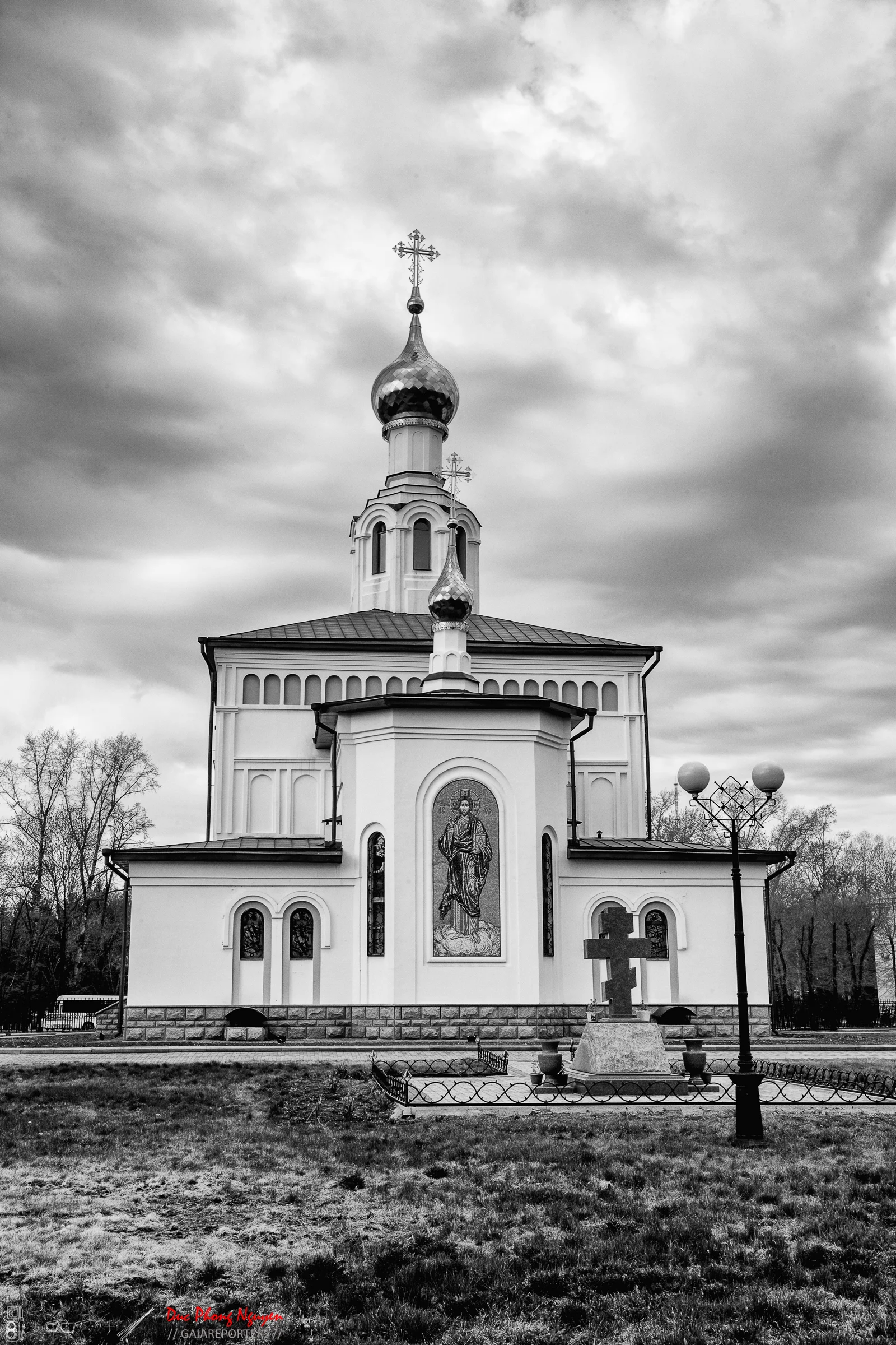 A black and white photo of a church with onion domes, an icon on the front, and a cross on top, in a grassy area with trees in the background and cloudy sky overhead.