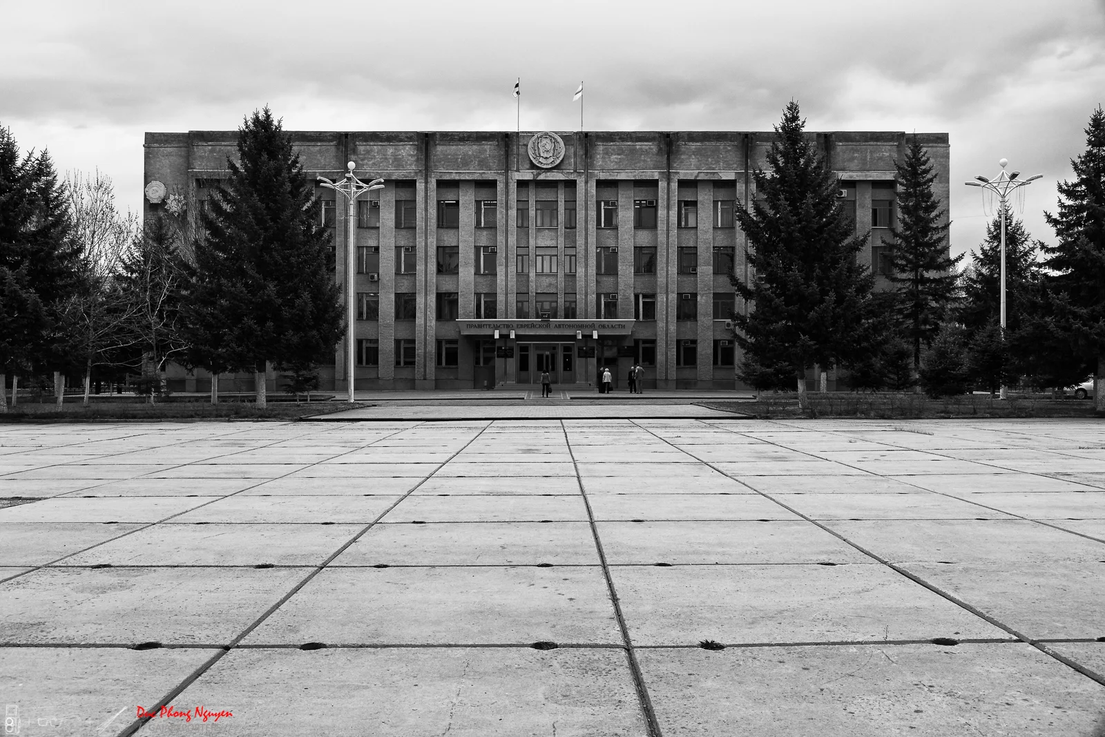 Black and white photo of a government building with flags on top, trees on both sides, and a paved open space in front with three people walking toward the entrance.