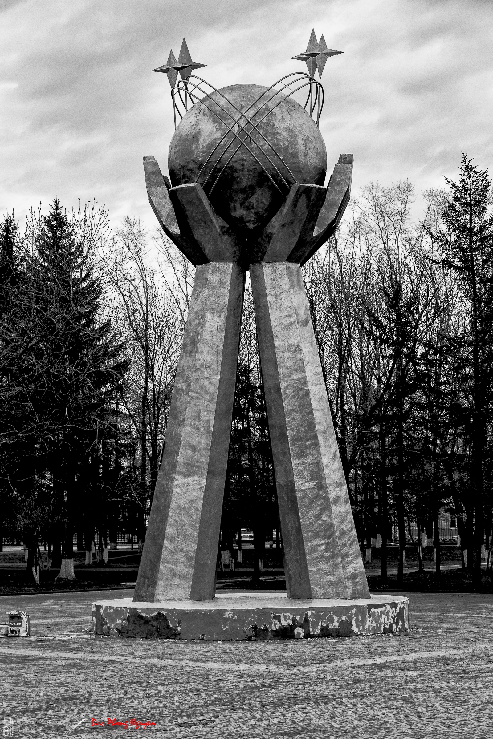 A tall sculpture with three large support beams, holding a sphere with metal wires and star-shaped adornments on top, set against a background of trees and cloudy sky.