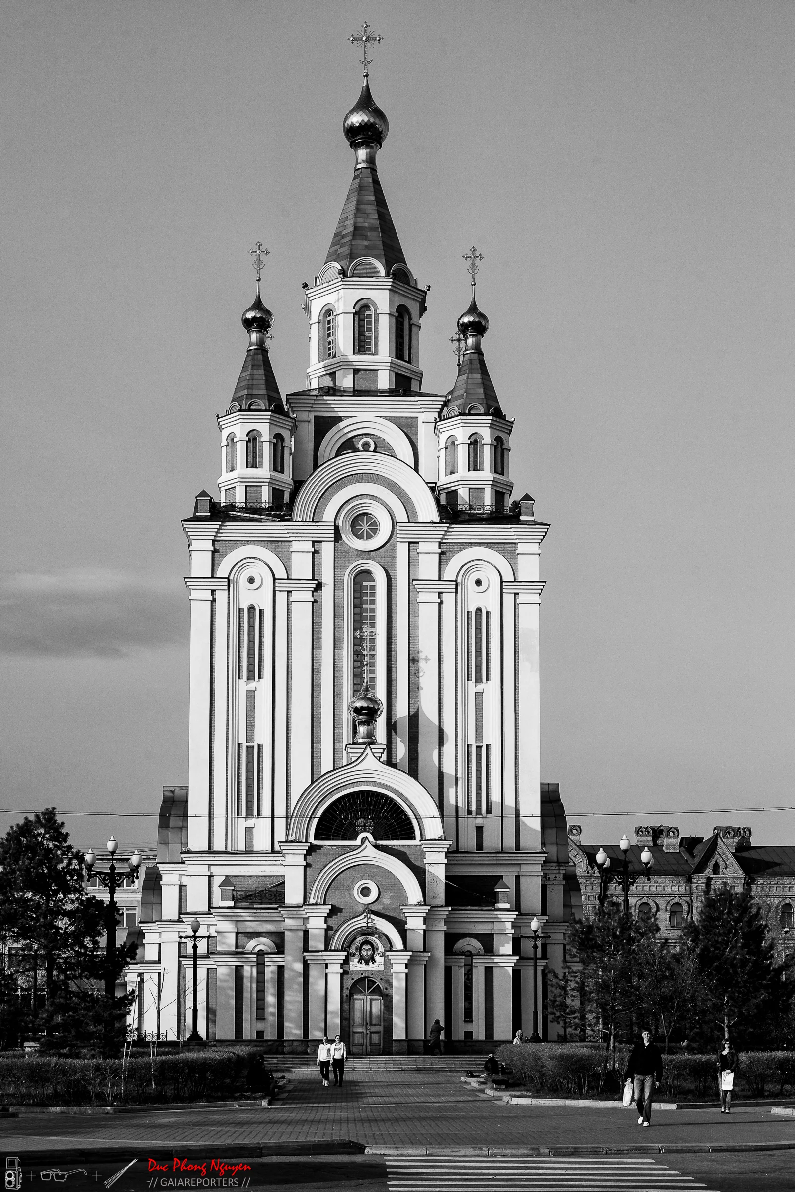 A black and white photo of a tall, ornate church with multiple domes and crosses on top, situated on a city street with people walking nearby.