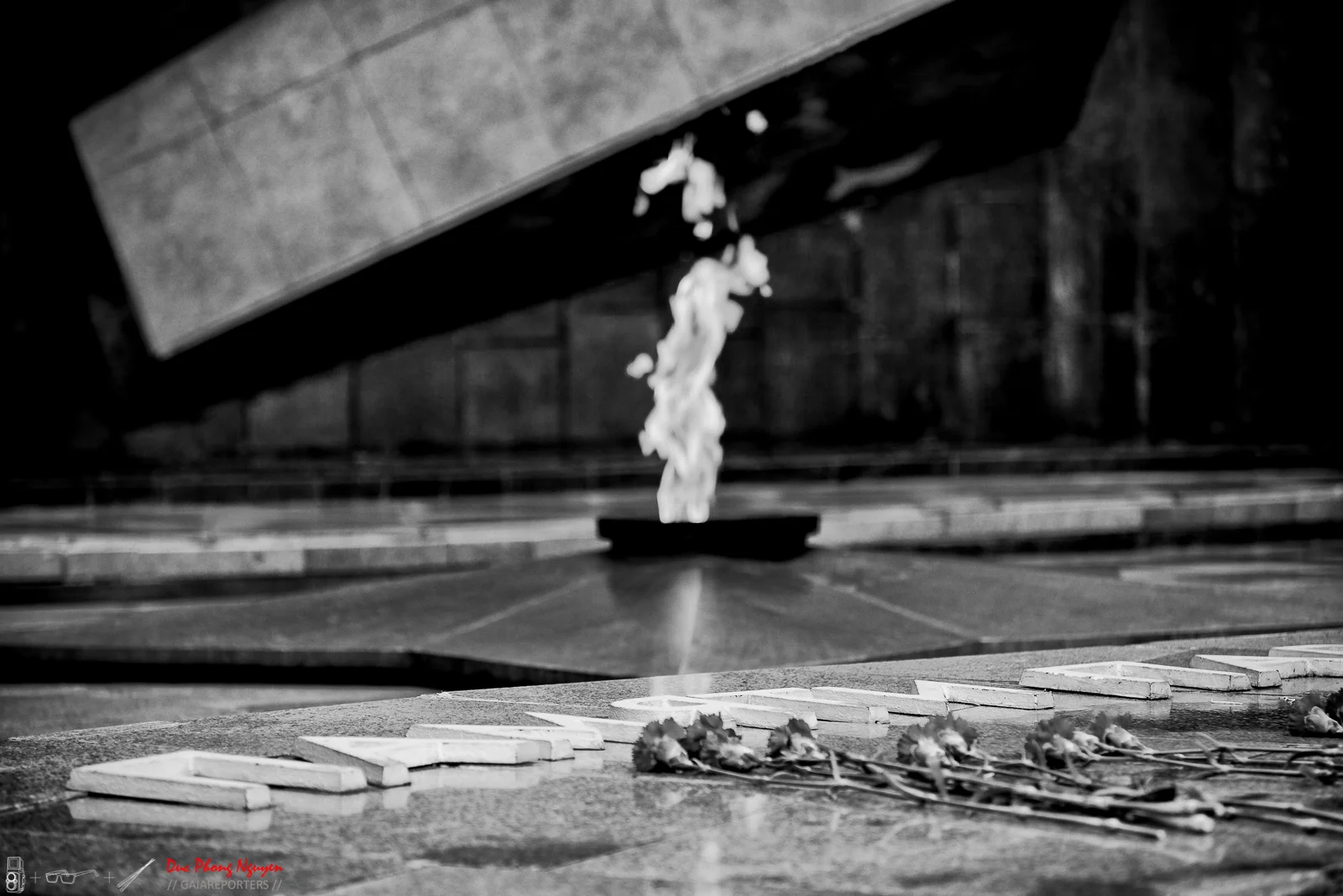 A black and white photograph of an eternal flame memorial with flowers laid in front, the flame burning in the center of a water feature with the word 'LOVE' spelled out with stones in the foreground.