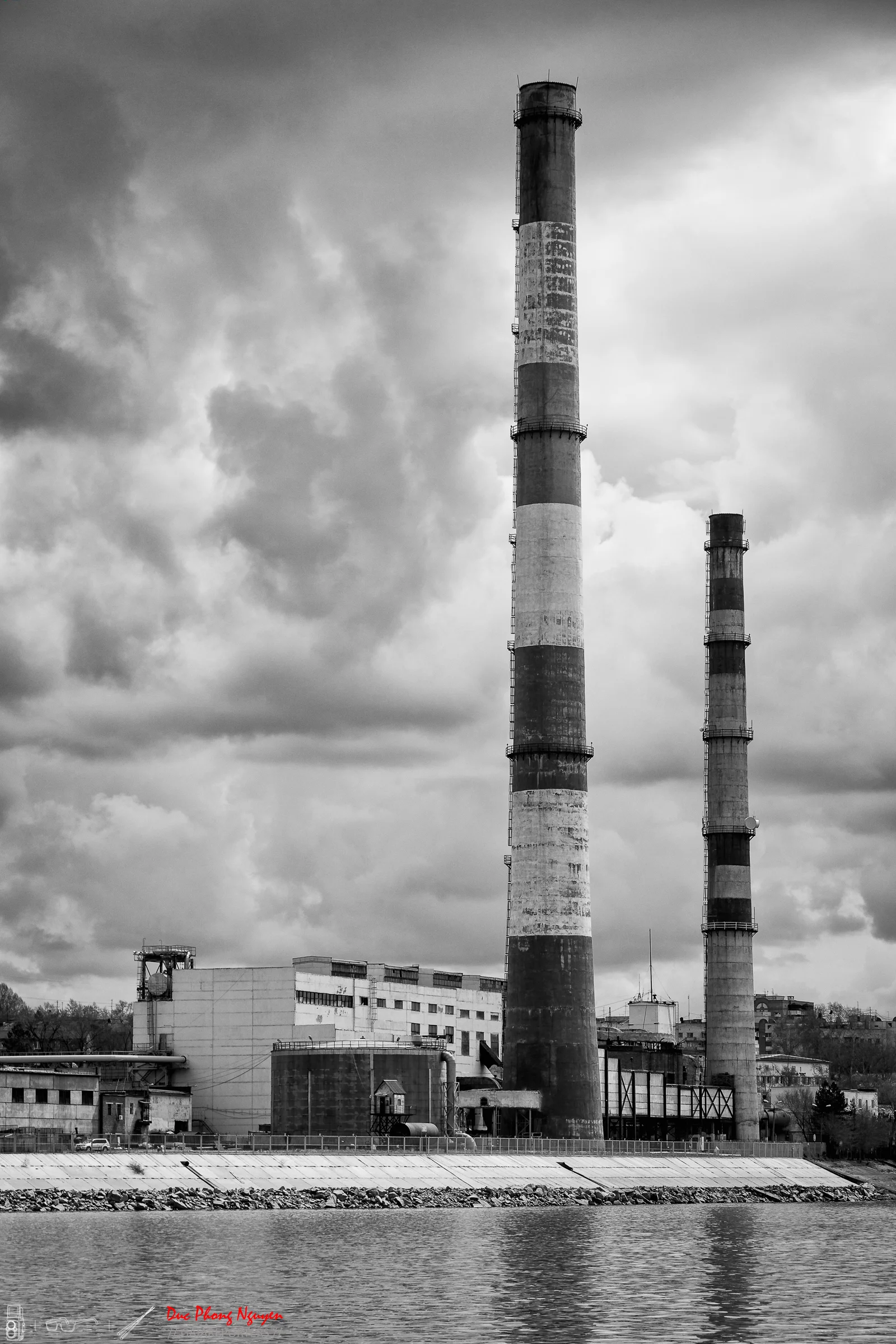 Black and white photo of an industrial factory with two tall smokestacks near a water body, under a cloudy sky.