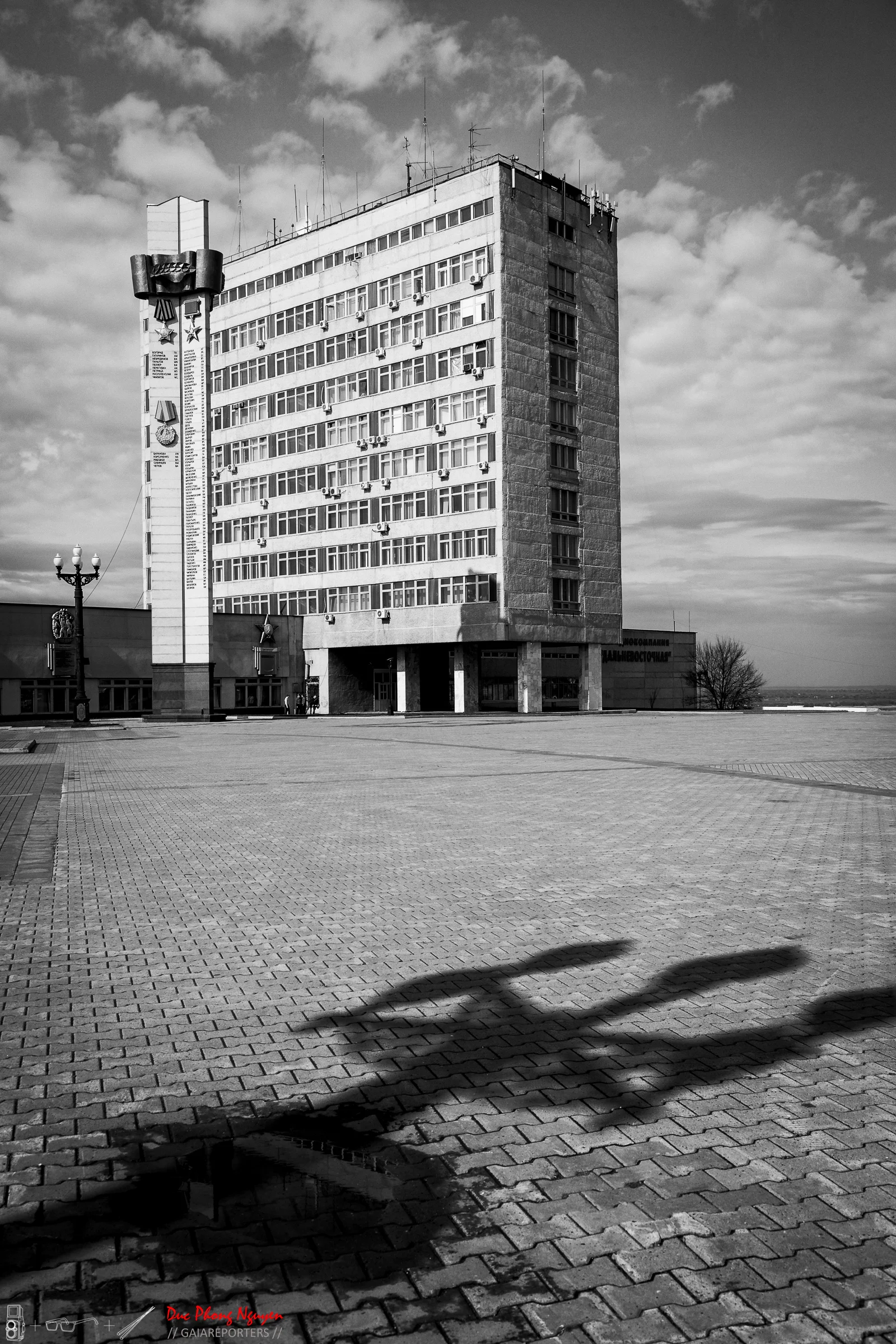 A black and white photo of a tall, multi-story building with rows of windows and air conditioning units, located on a paved open space with a street lamp and shadows cast on the ground.