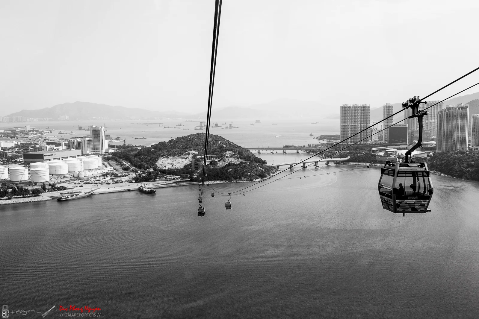 A black and white photo of a cityscape viewed from a cable car ride over water. The city has tall buildings and mountains in the background, with oil tanks visible near the shore.