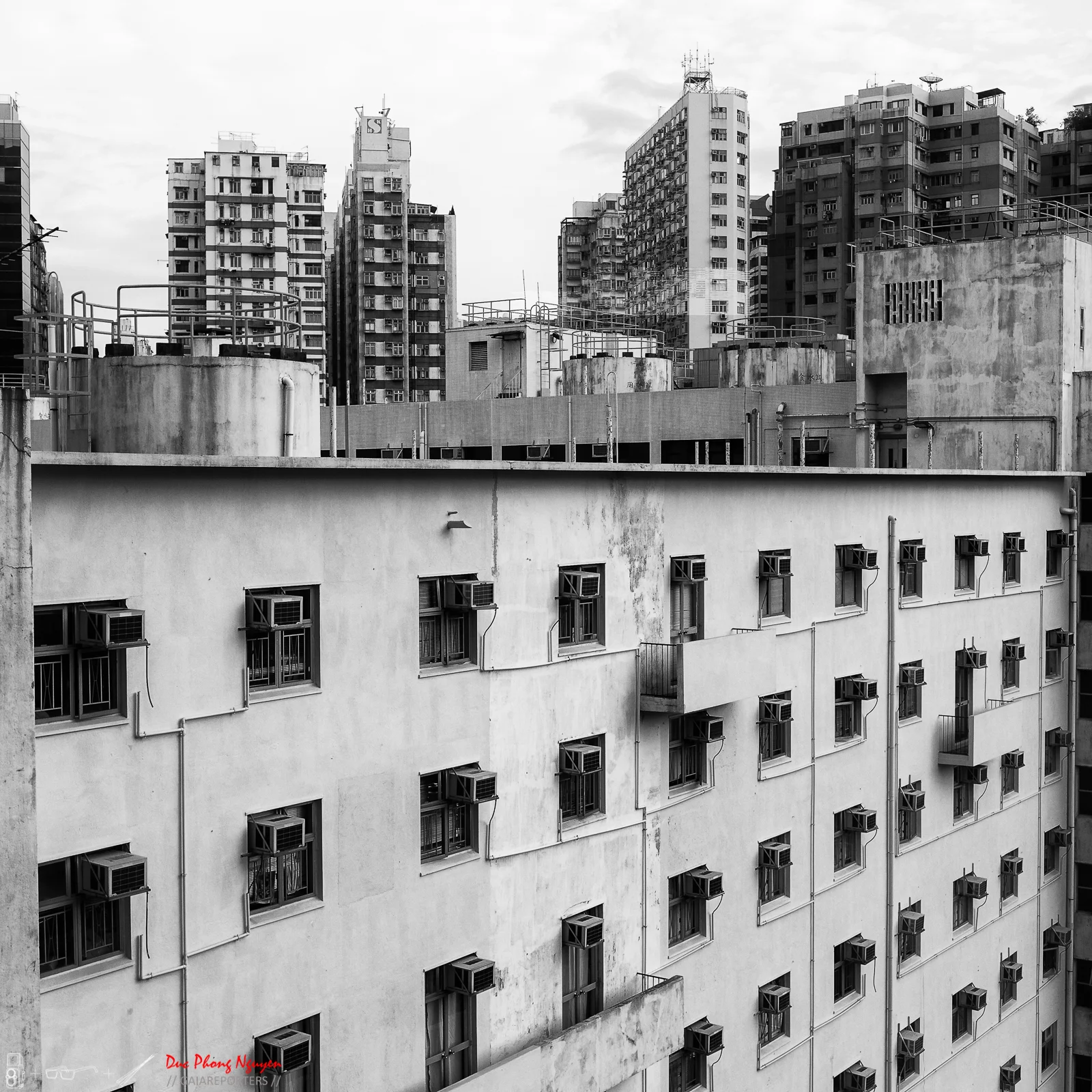 Black and white photo of a cityscape with numerous apartment buildings, some with air conditioning units attached to the windows, and taller skyscrapers in the background.