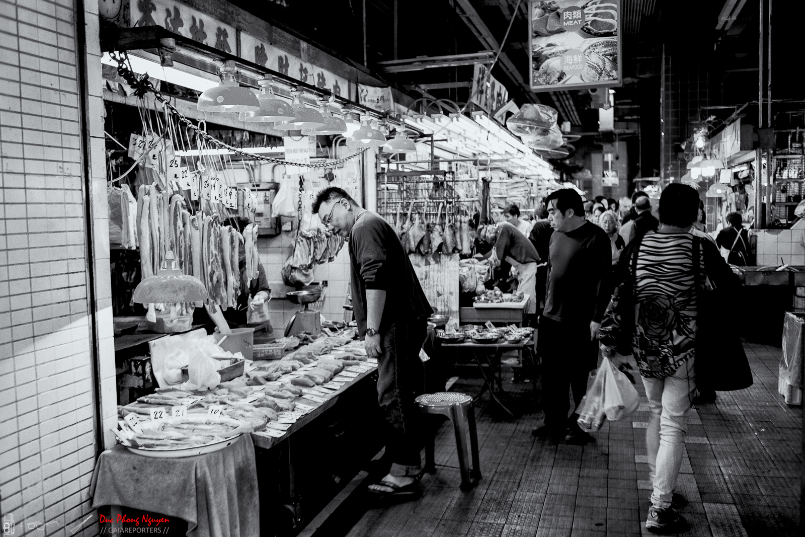 A busy indoor market with people shopping for meat and seafood. A man is inspecting meat on a counter, and other shoppers are walking and browsing. Hanging meats and seafood are displayed, and signs with prices are visible.