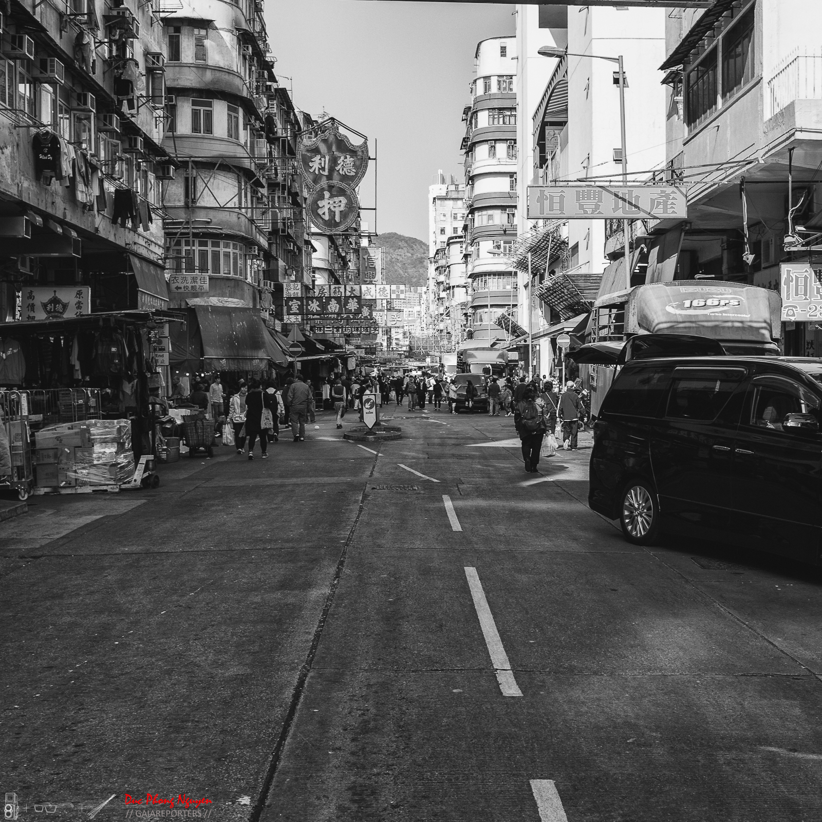 A busy city street scene in Hong Kong with pedestrians walking and shopping, surrounded by tall buildings with Chinese signage and street vendors.