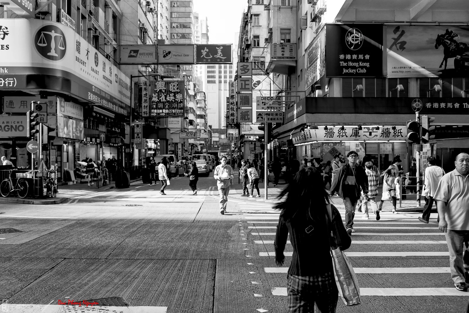 Black and white photo of a busy city street intersection with pedestrians crossing. Buildings are lined with signs and advertisements in Chinese characters. Vehicles are parked along the street, and streetlights and traffic signals are visible.