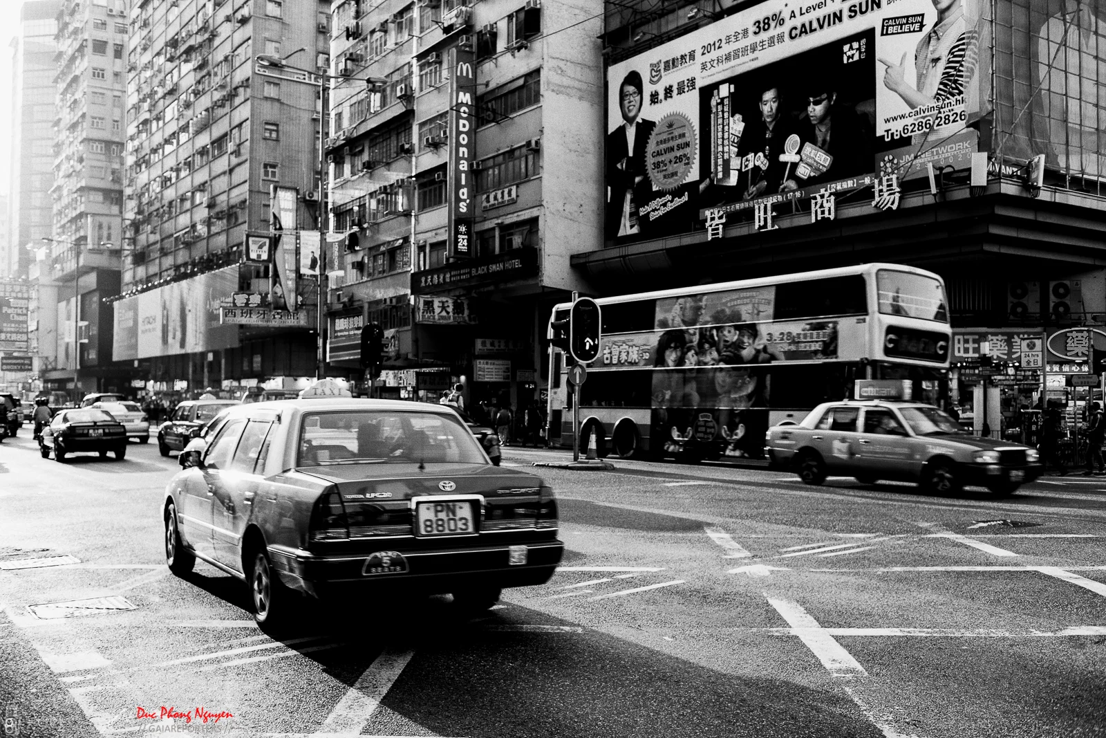 Black and white street scene with cars, taxis, and a double-decker bus on an urban road. Tall buildings with numerous advertisements and billboards line the street. Pedestrians are visible on the sidewalks, and the area has a busy city atmosphere.