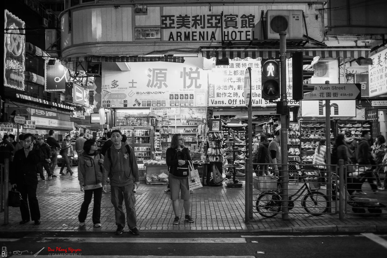 Black and white photo of a busy street scene in an Asian city, with pedestrians waiting at a crosswalk, shops, and signs in Chinese and English, including a sign for Armenia Hotel.