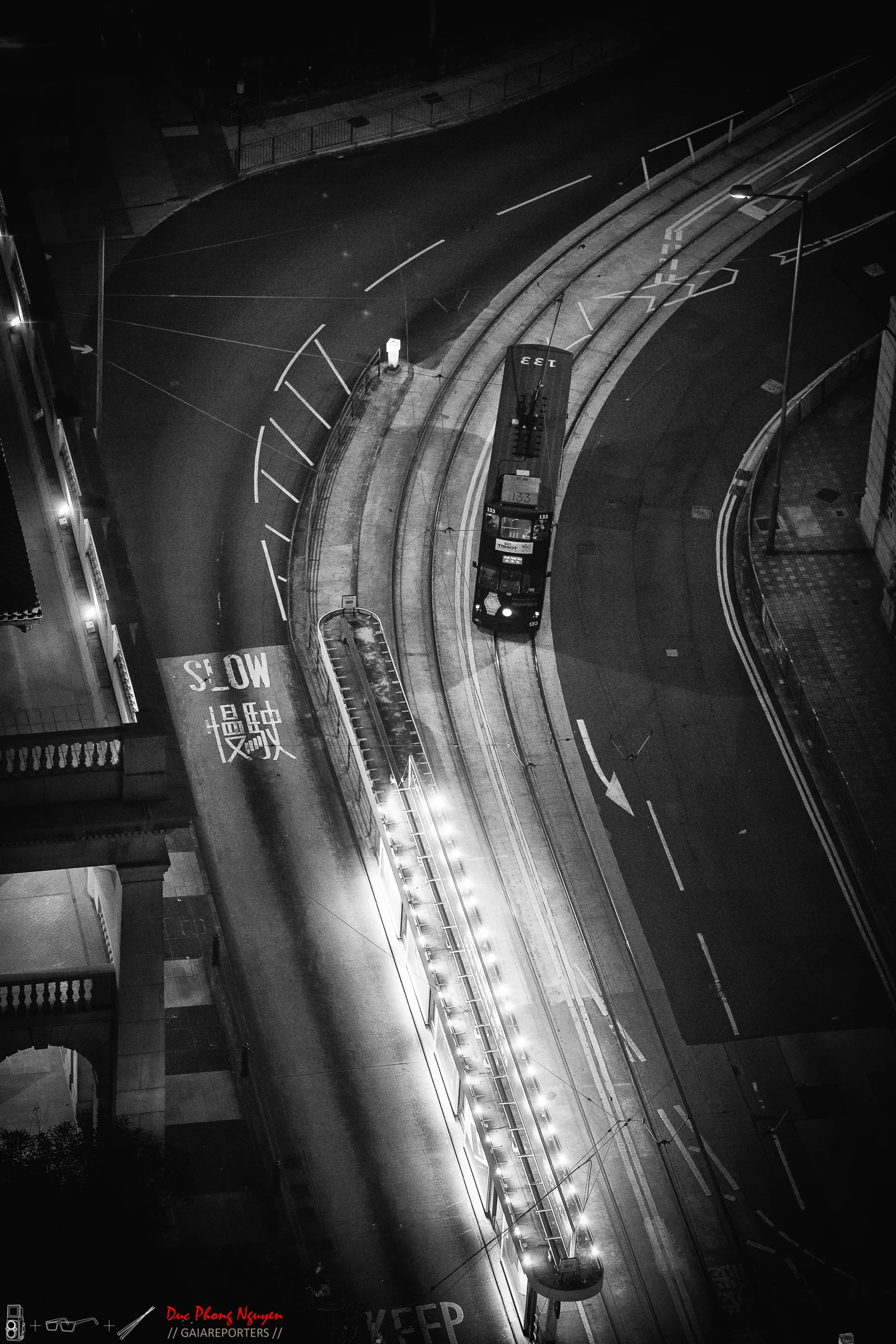 A black-and-white photo of a city street at night showing an empty bus on tram tracks, street lights, and a curved road with markings, including a "SLOW" sign in Chinese characters.