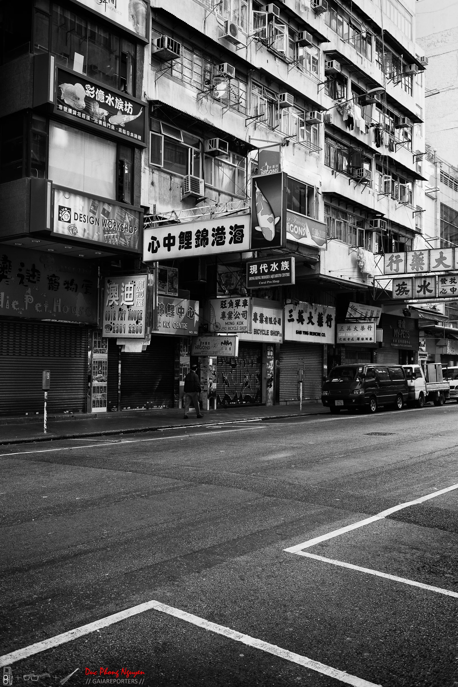 Black and white photo of a city street with shops and residential buildings. Storefront signs display Chinese characters and some English words, and a few parked vehicles are visible. A person is walking on the sidewalk.