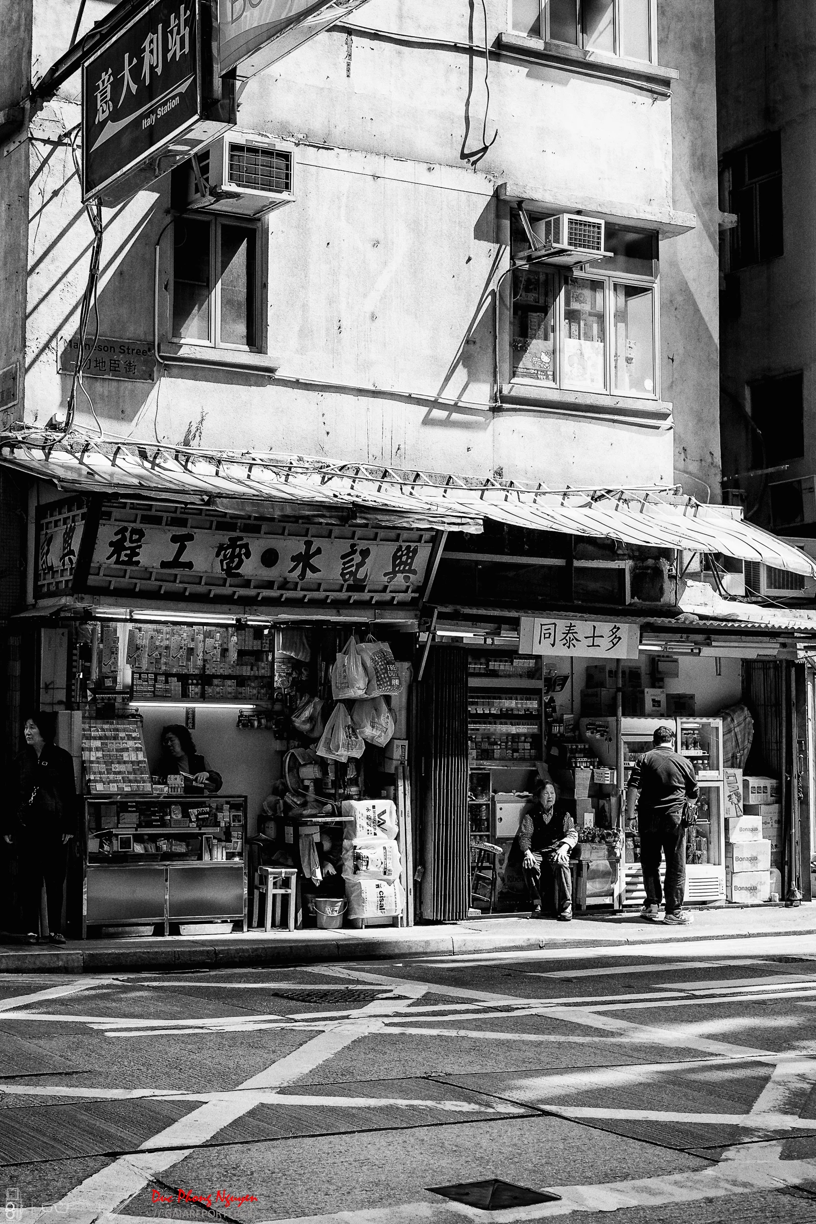 A black and white photo of a street corner with a small shop selling various items, two women sitting outside, one man standing, and a building with windows and air conditioning units above the shop.