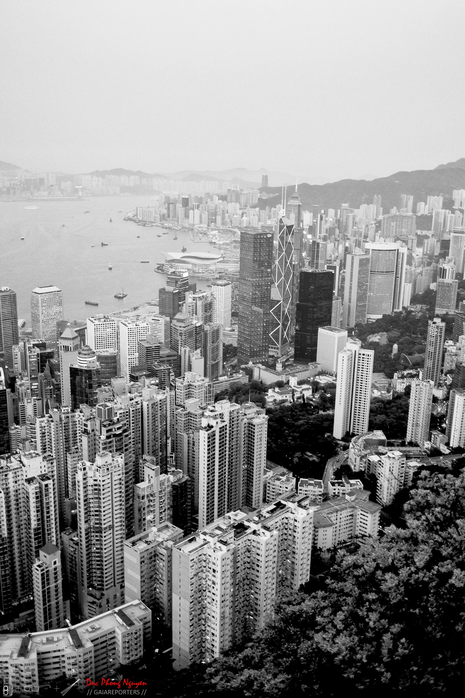 Black and white aerial view of Hong Kong skyline with dense skyscrapers, Victoria Harbour, and surrounding mountains.