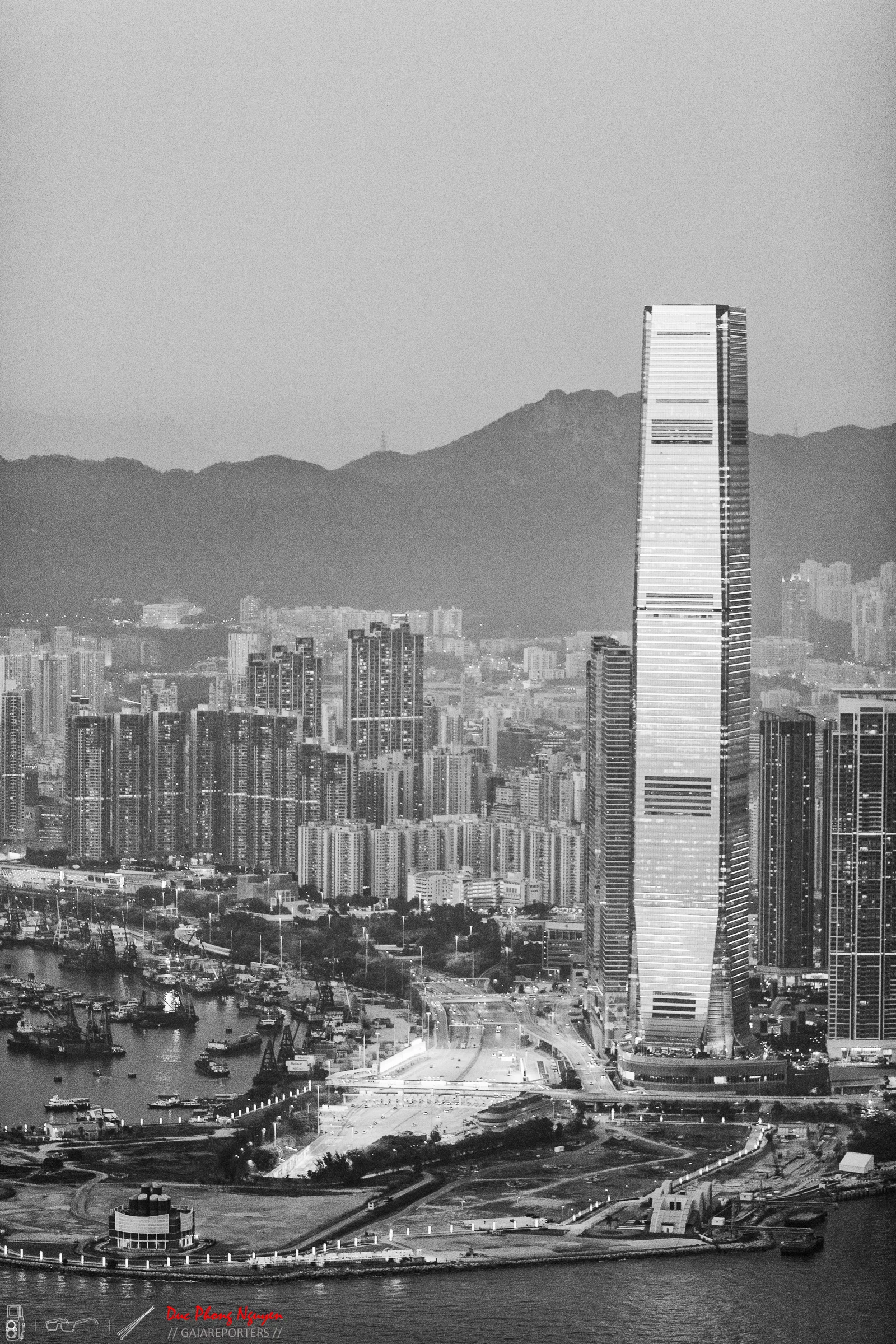 Black and white photograph of a city skyline featuring a tall, modern skyscraper, with a river and boats in the foreground, and mountains in the background.