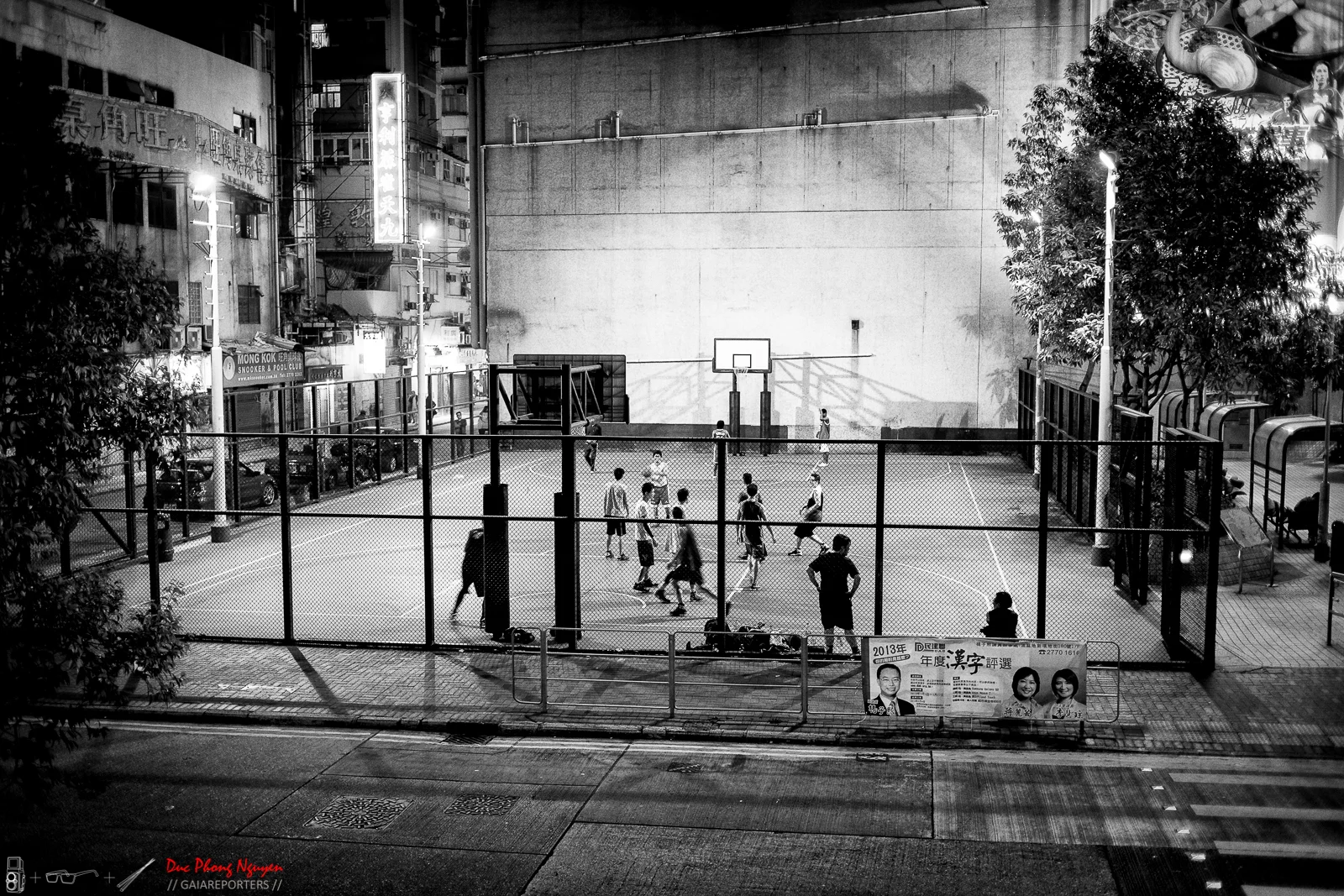 Nighttime black and white photograph of an outdoor basketball court with children playing. The court is enclosed by a chain-link fence and surrounded by urban buildings, trees, and streetlamps.