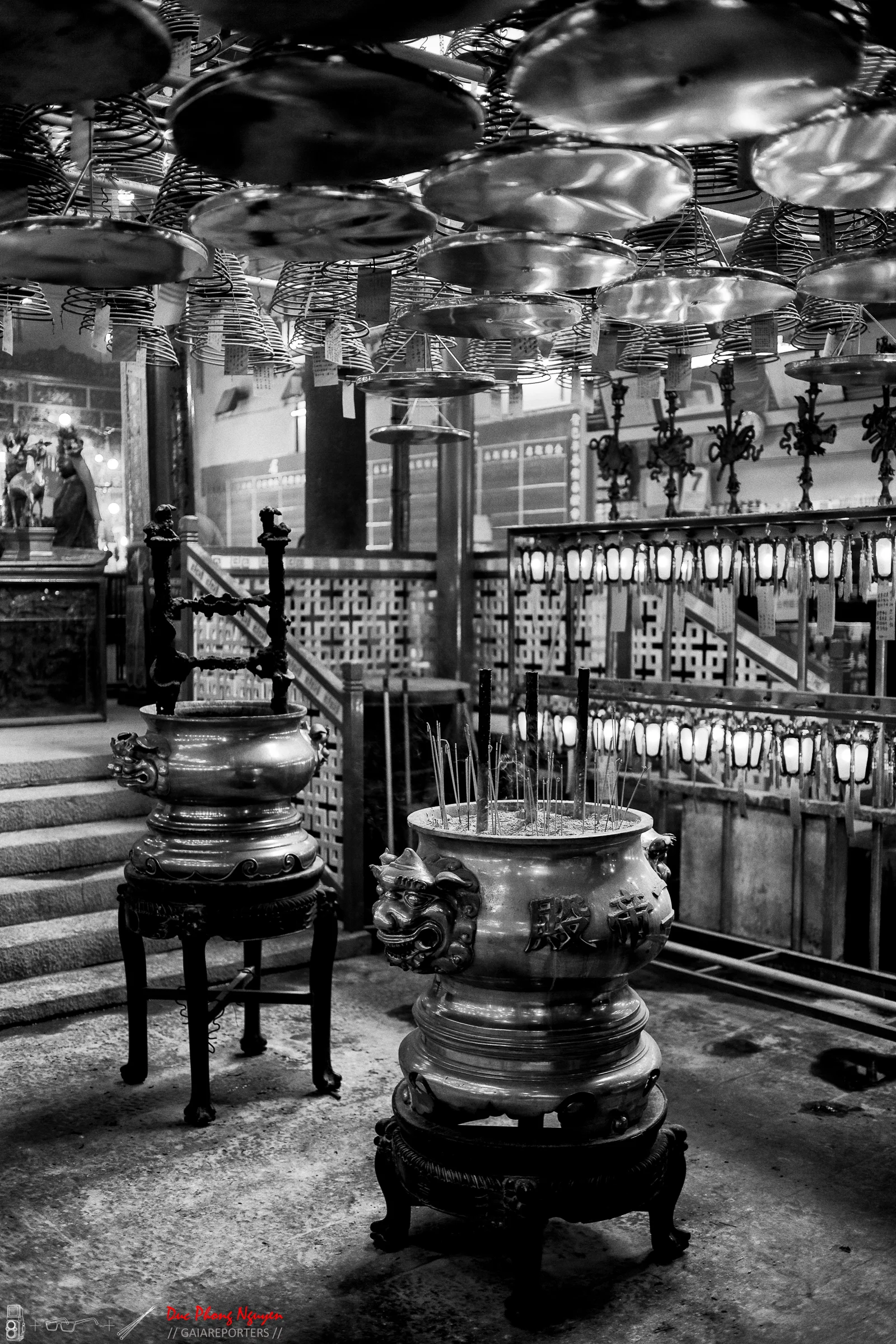 Black and white photo of incense burners with incense sticks inside, hanging plates above, and lanterns on the walls inside a temple or shrine.