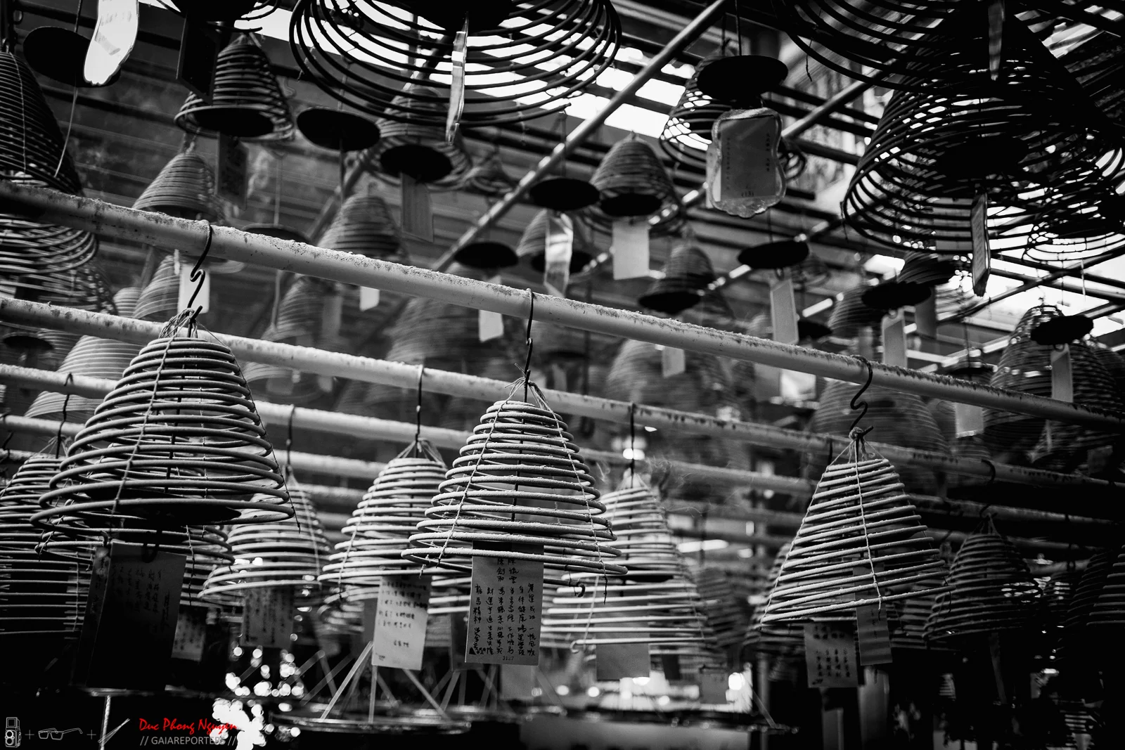 Black and white photo of various woven bamboo or rattan lampshades hanging from a ceiling in a market or shop.