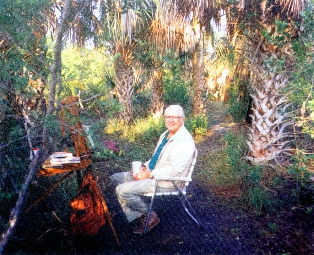 William North, an American painter, with glasses sitting in a white metal chair in a lush, green outdoor Florida garden, with trees and plants surrounding him, holding a white cup, and a wooden desk with papers and a reading lamp nearby.