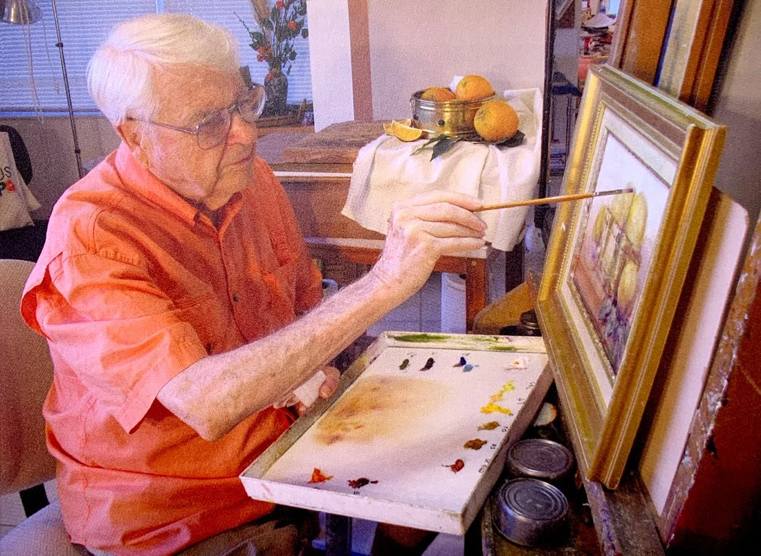 An elderly man with white hair and glasses painting on a canvas, using a paintbrush. He is seated at a table with a paint palette and is working on a framed picture. In the background, there is a table with lemons and a flower arrangement.