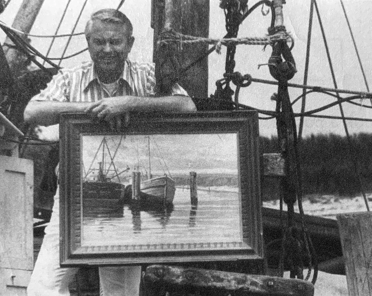 William North, an American painter, with light hair smiling, holding a framed painting of boats in the water, in an outdoor setting with a large boat and rigging in the background.