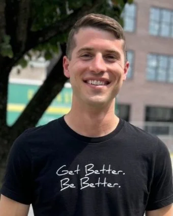 Young man outdoors smiling, wearing a black t-shirt with the text 'Get Better. Be Better.'