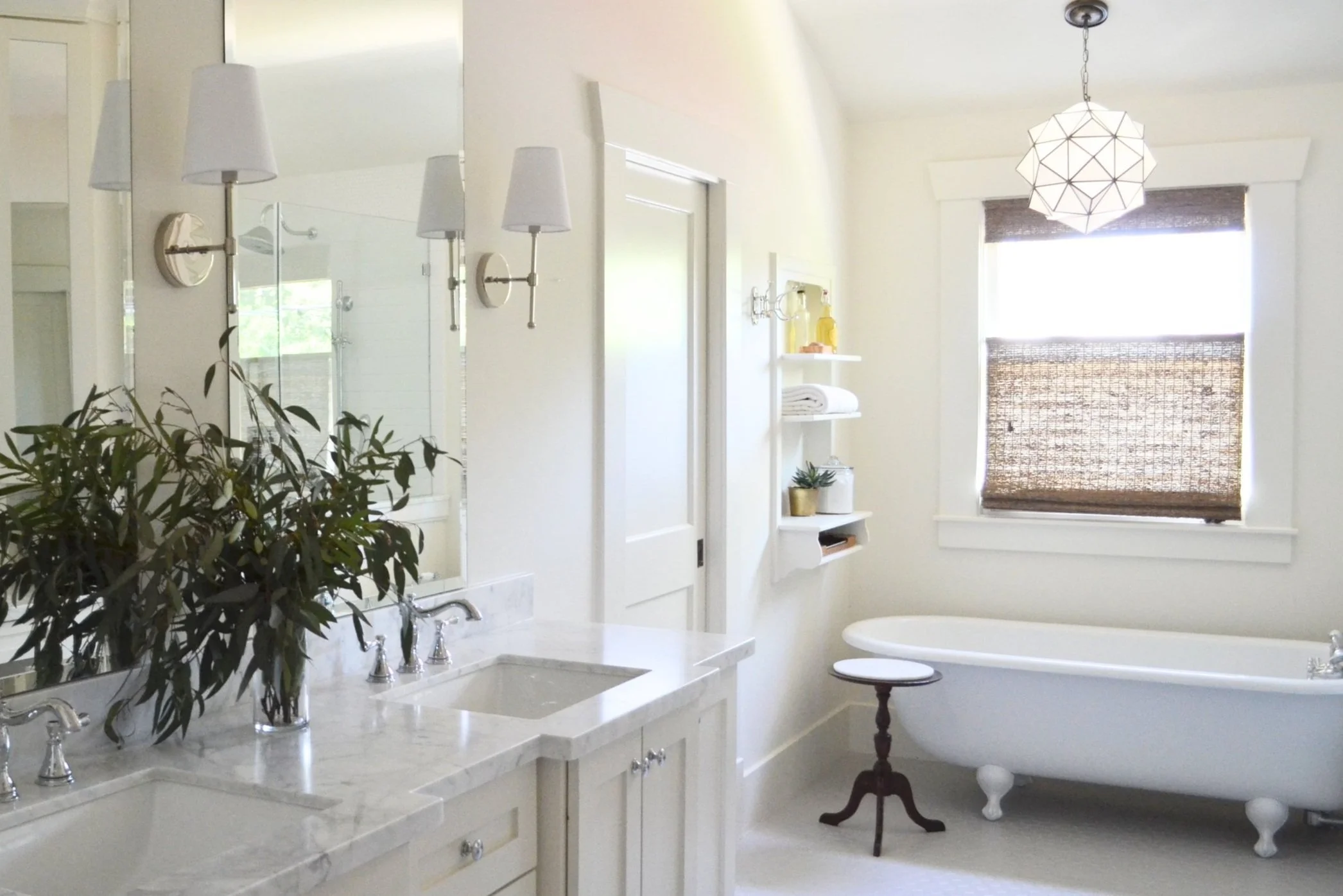 A modern bathroom with a marble-topped double sink vanity, a large mirror, wall-mounted lights, and a potted plant. A freestanding bathtub is near a window with woven blinds. A geometric pendant light hangs from the ceiling, and a small round table sits beside the tub.