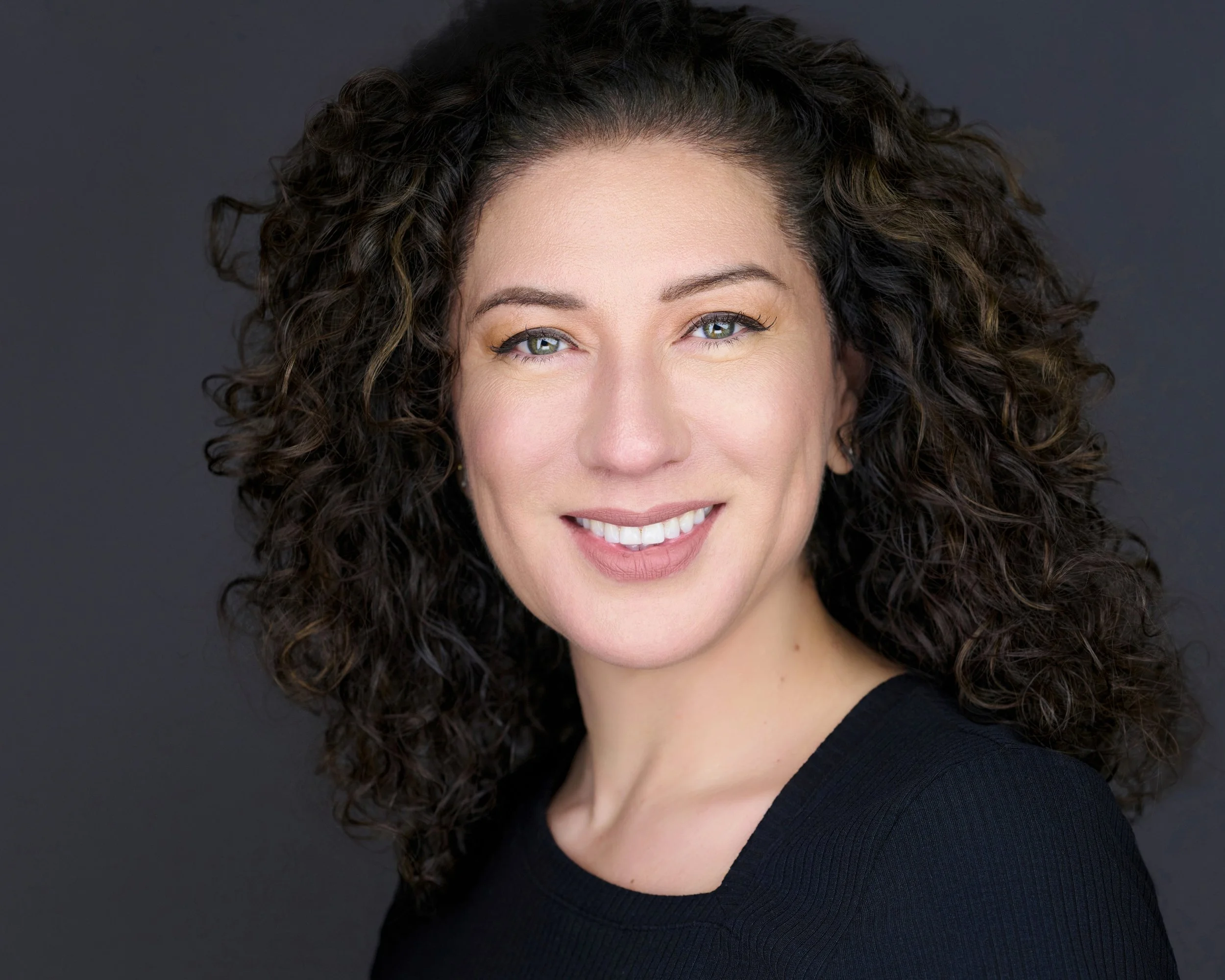 Close-up of a woman with curly brown hair and blue eyes, smiling, wearing a dark top against a dark background.