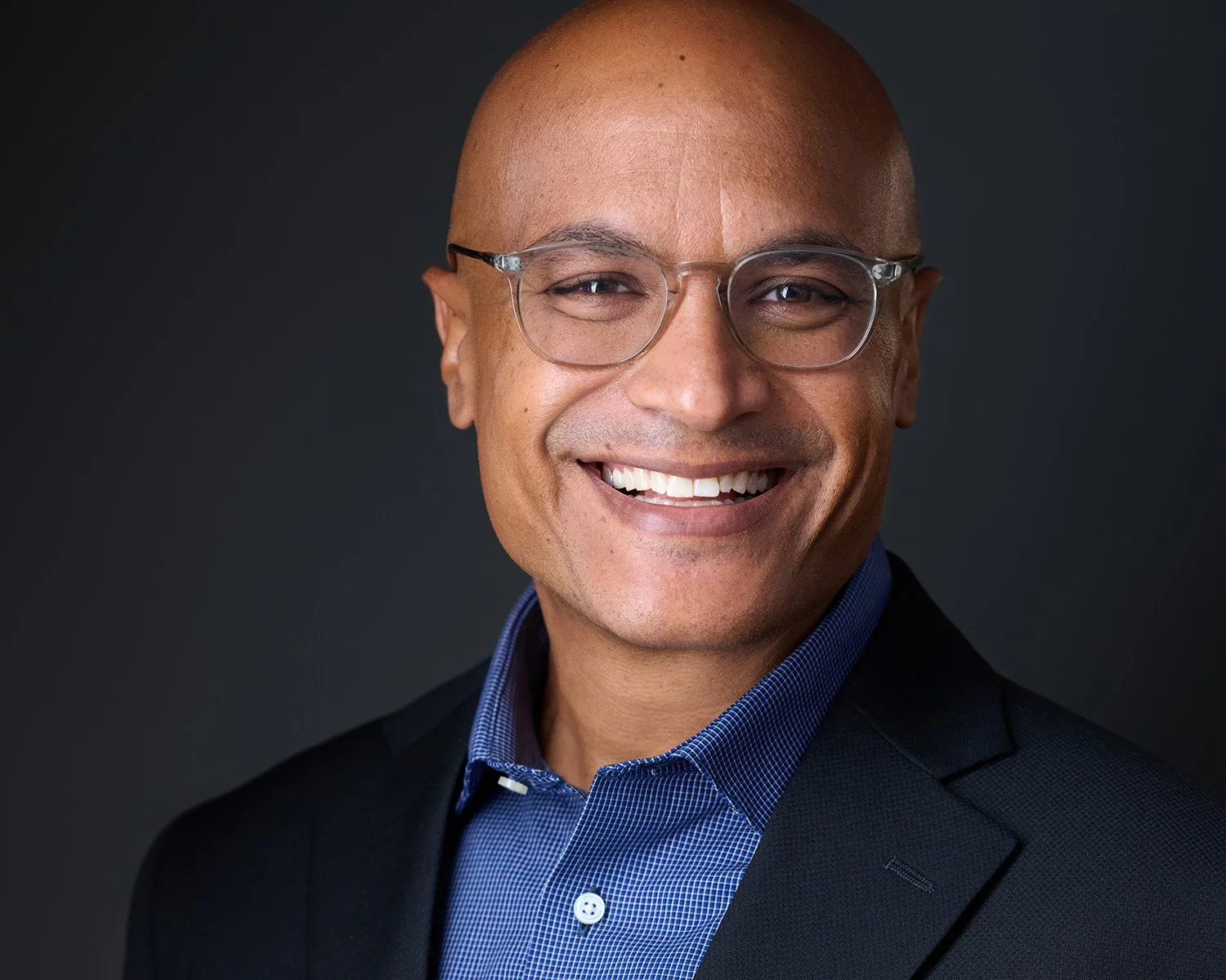 Studio Headshot of a Bald Man in a Dark Suit with a Blue Shirt wearing glasses with a smile