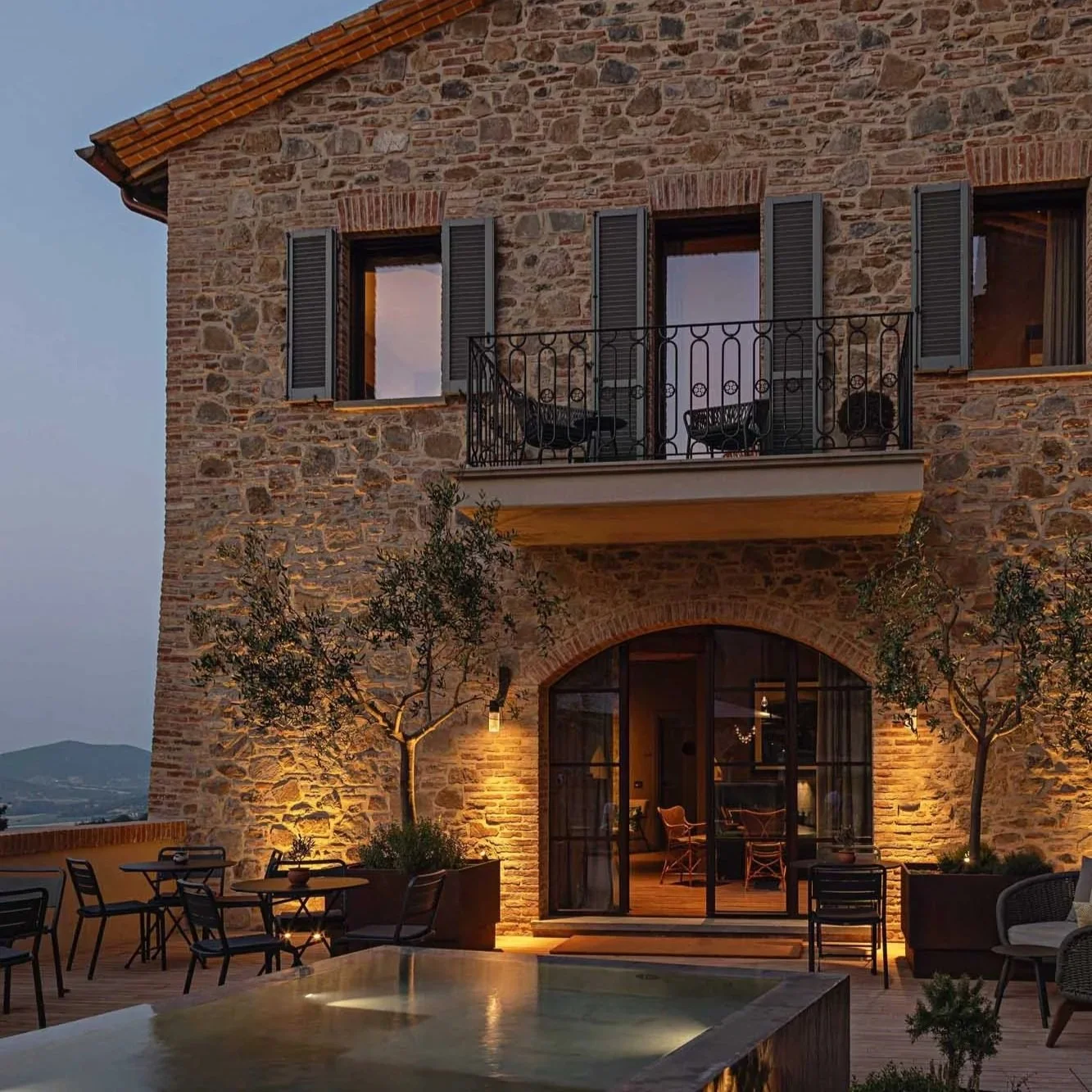 A multi-story stone house with open windows and a small balcony with chairs. The house has warm lighting and is surrounded by potted trees and outdoor furniture, creating a cozy atmosphere at dusk.