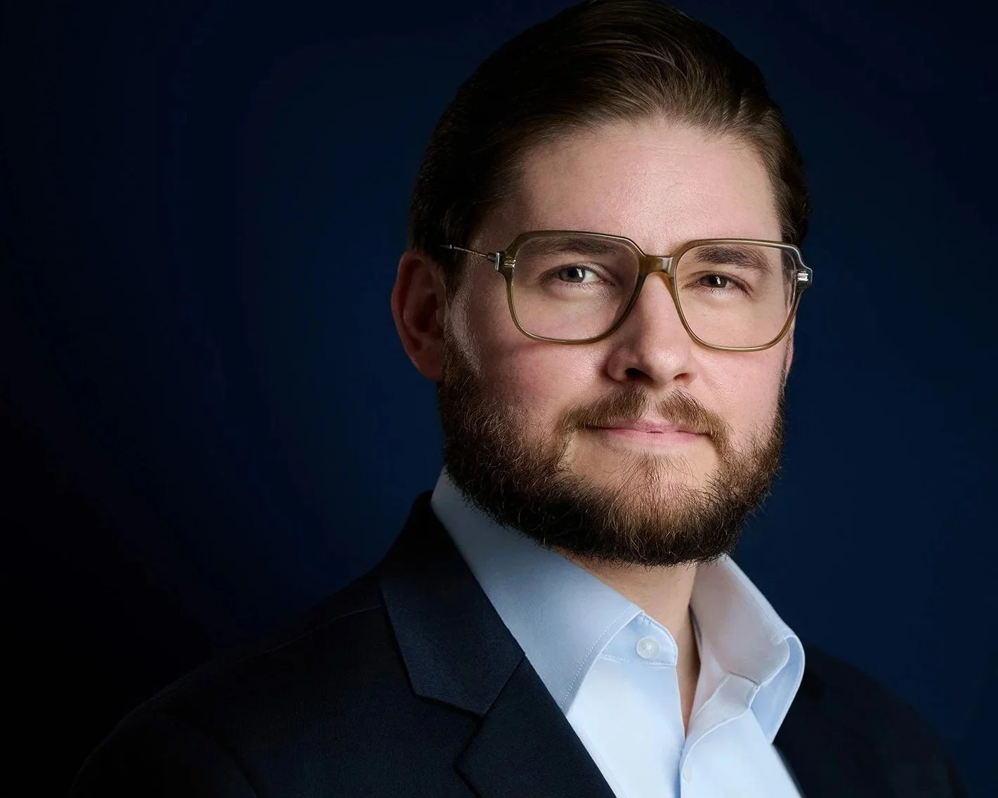 A young man with glasses and a beard wearing a dark suit and white shirt, posing against a dark background.
