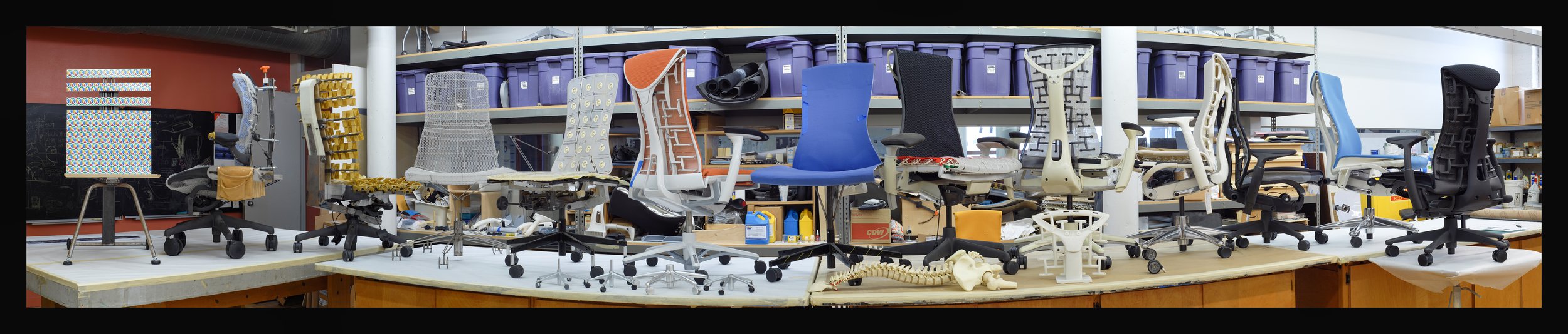 Collection of ergonomic office chairs in various colors and styles on a work table in a workshop setting.