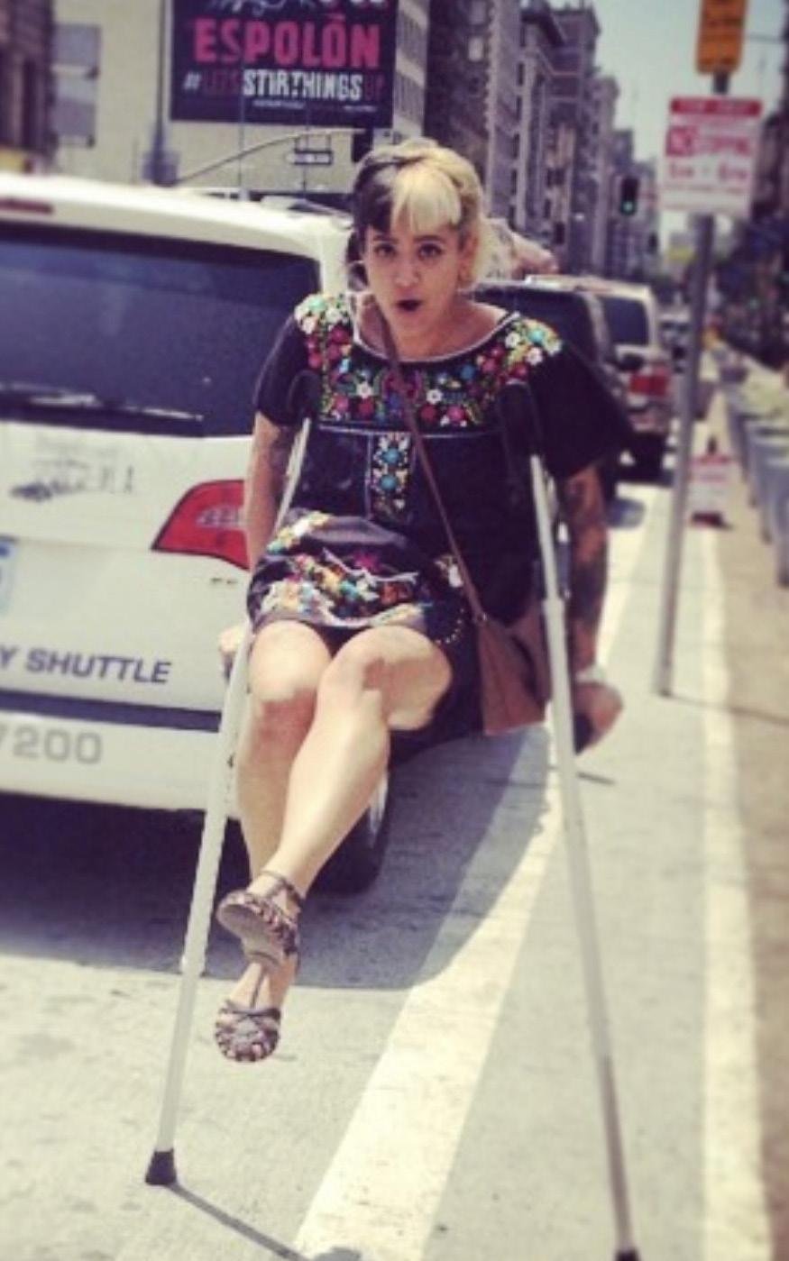A woman with short blonde hair, sitting on a wheelchair, is surprised or alarmed, on a city sidewalk with cars and buildings in the background. She is wearing a black dress with colorful floral embroidery and high-heeled sandals.
