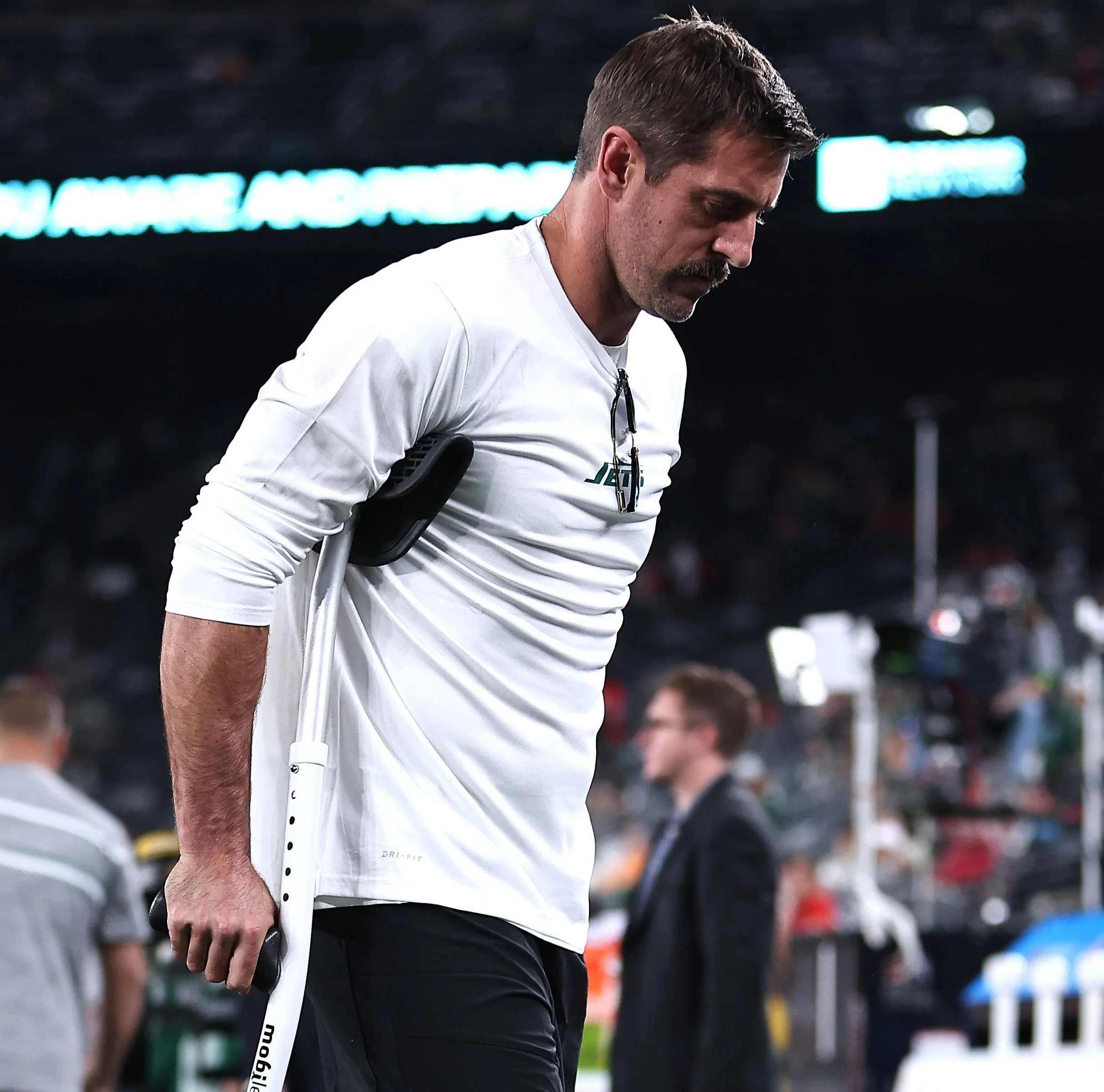 A man with a mustache and brown hair, wearing a white shirt, holding a crutch, looking down during a sporting event, with a blurred crowd in the background.