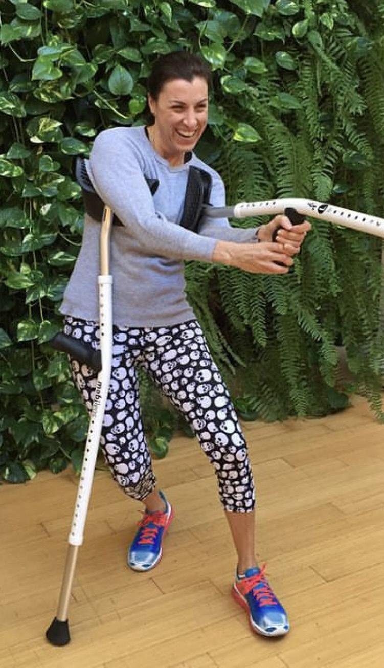 Woman with a prosthetic leg participating in adaptive fitness, holding a white stick, smiling, standing on a wooden floor in front of a leafy green background.