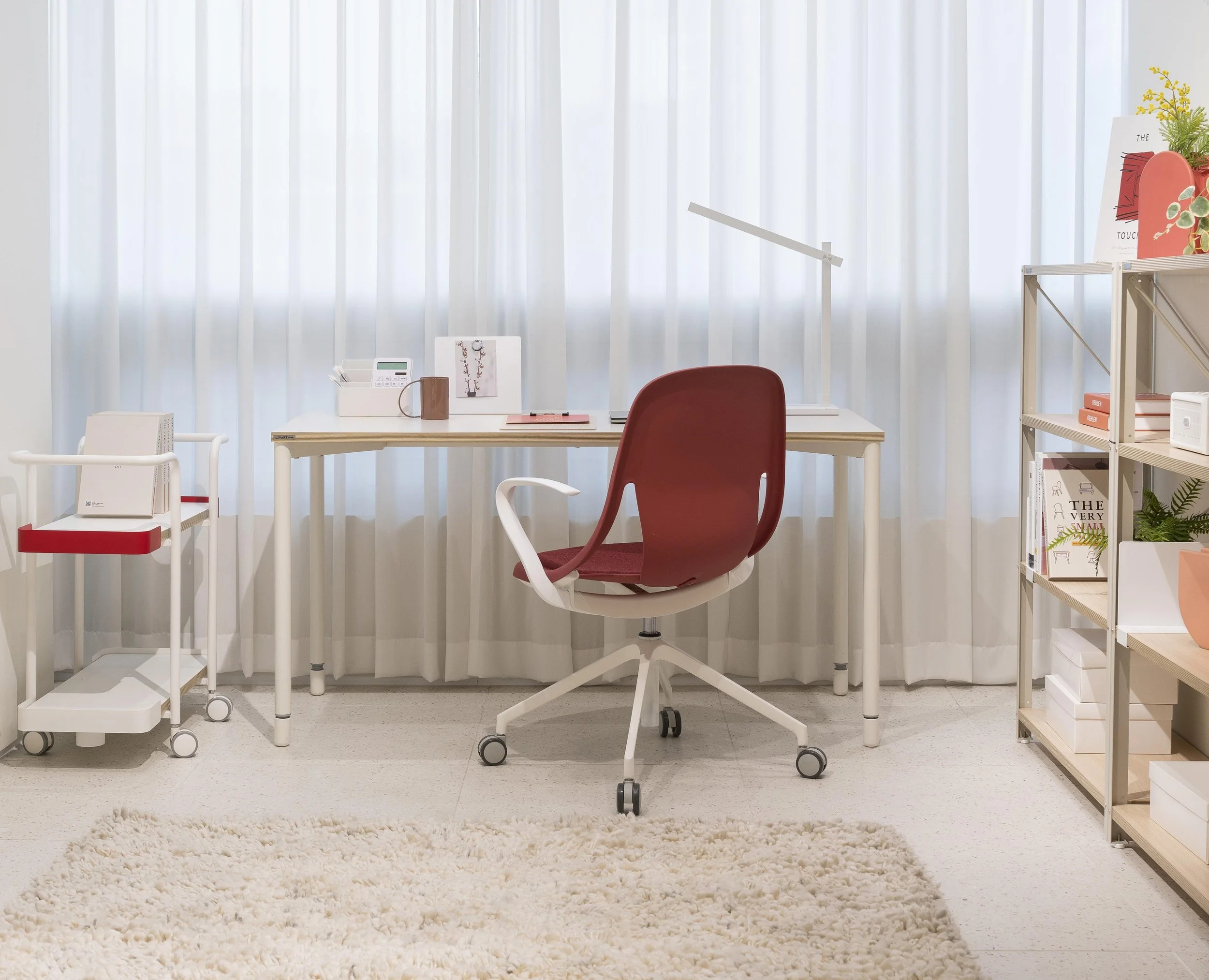 A home office space with a white desk, red and white chairs, bookshelf, and decorative items including a plant and books, in front of white curtains.