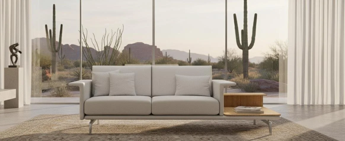 Modern living room with large windows showing desert landscape with cacti, featuring a white sofa, side table with books, curtains, and desert scenery in the background.