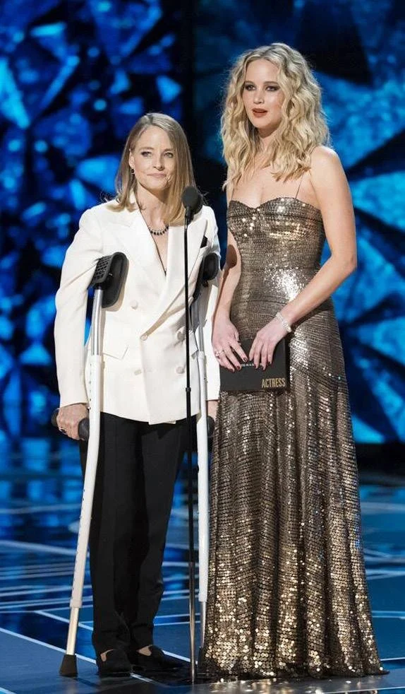 Two women standing on a stage. The woman on the left has crutches and is dressed in a white blazer and black pants. The woman on the right is wearing a gold, shiny, strapless gown and holding a black award envelope labeled 'ACTRESS.'