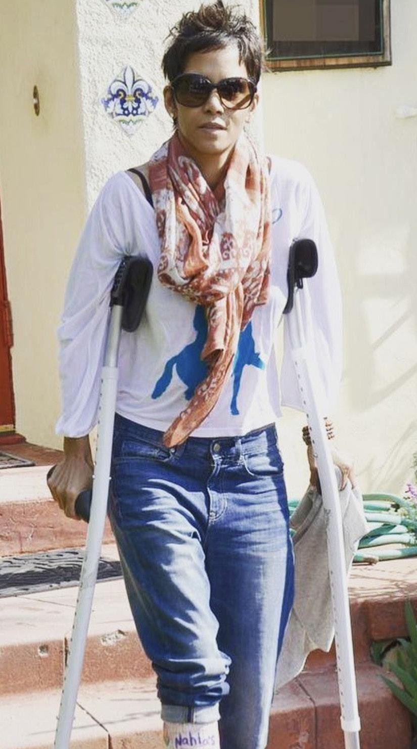 A woman with short dark hair wearing sunglasses, a pink scarf, a white long-sleeve shirt, and blue jeans, walking with crutches outside a house.