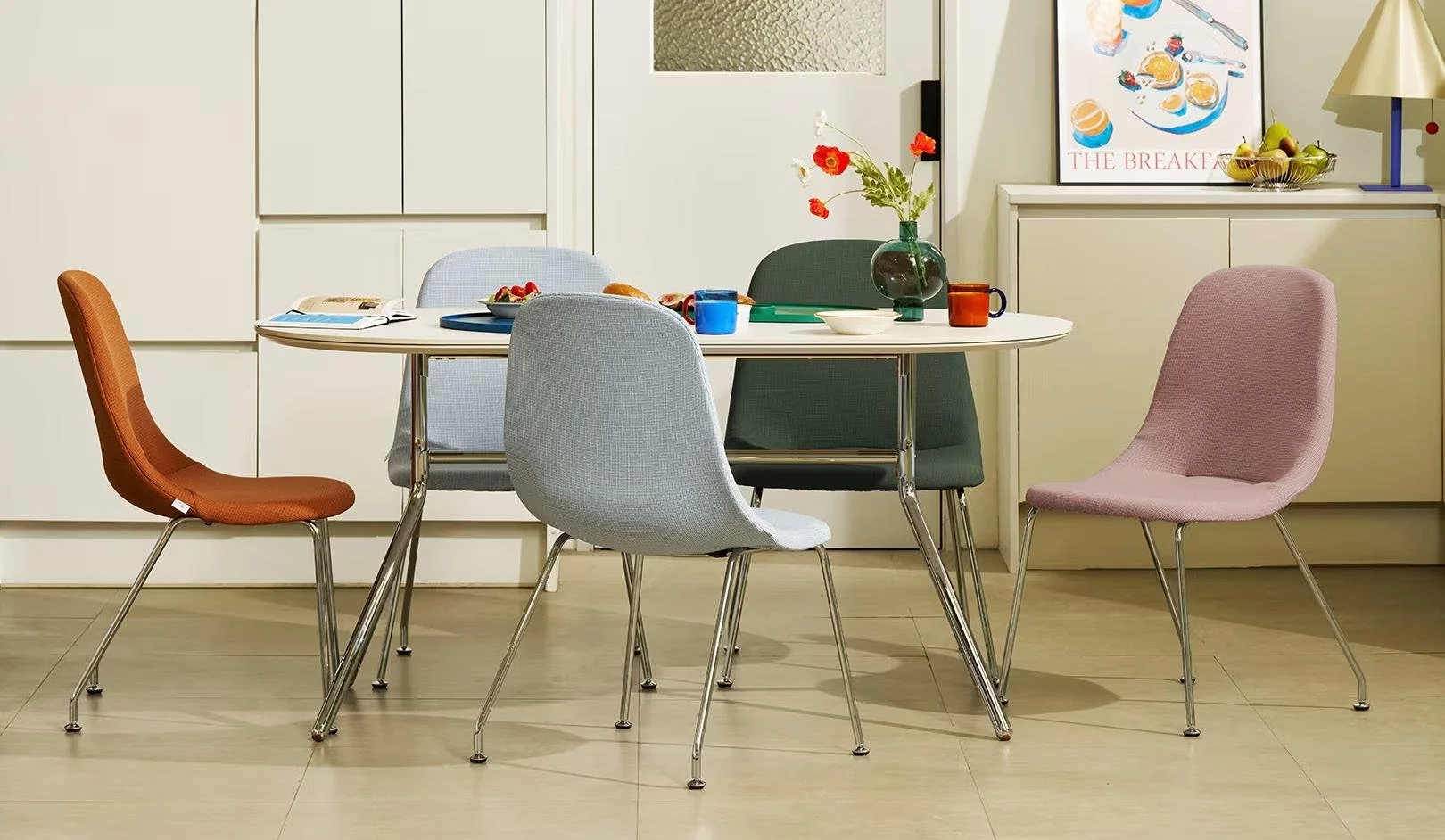 A dining room with a white oval table surrounded by five colorful chairs, set with breakfast items, a glass vase with red flowers, and a framed picture on a cabinet. The room has light-colored walls and wooden flooring.